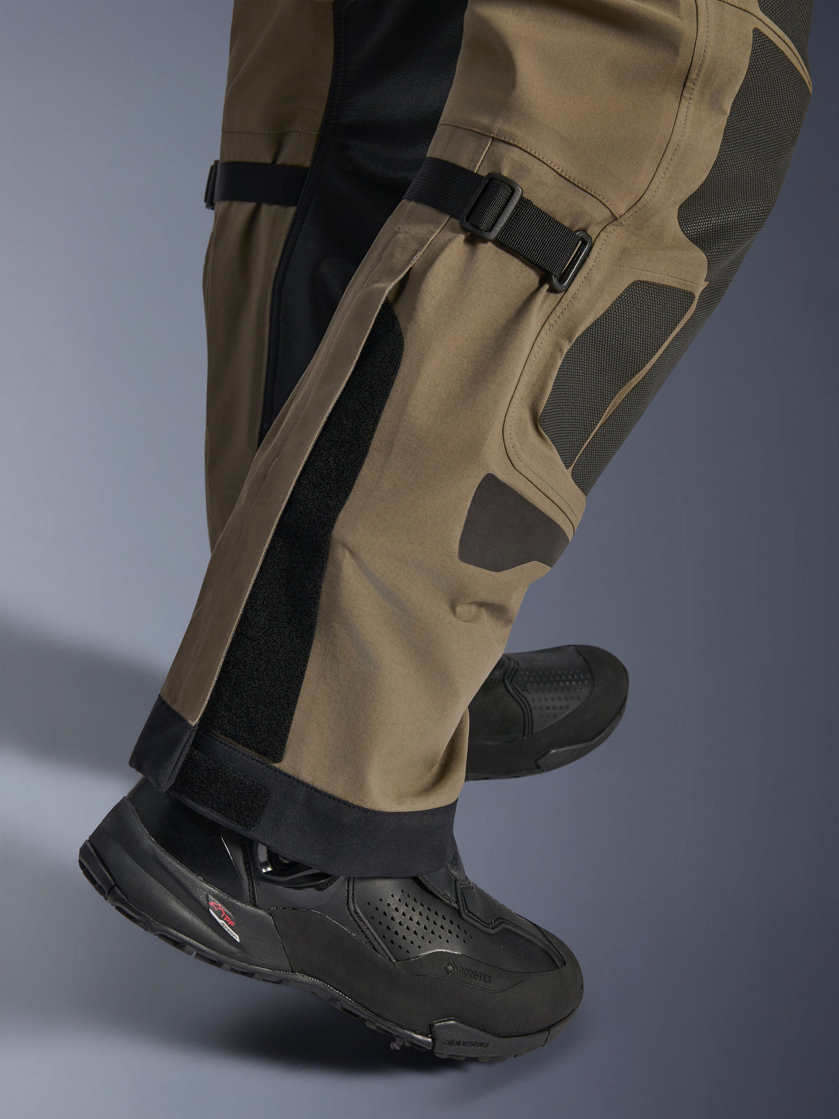 Close-up van Alpinestars Nazca 3L GORE-TEX PRO Broek in Walnut Black, met technische details van het onderbeen inclusief kuitvolume-verstellers en Superfabric-versterkingen, gedragen met zwarte adventure motorlaarzen.