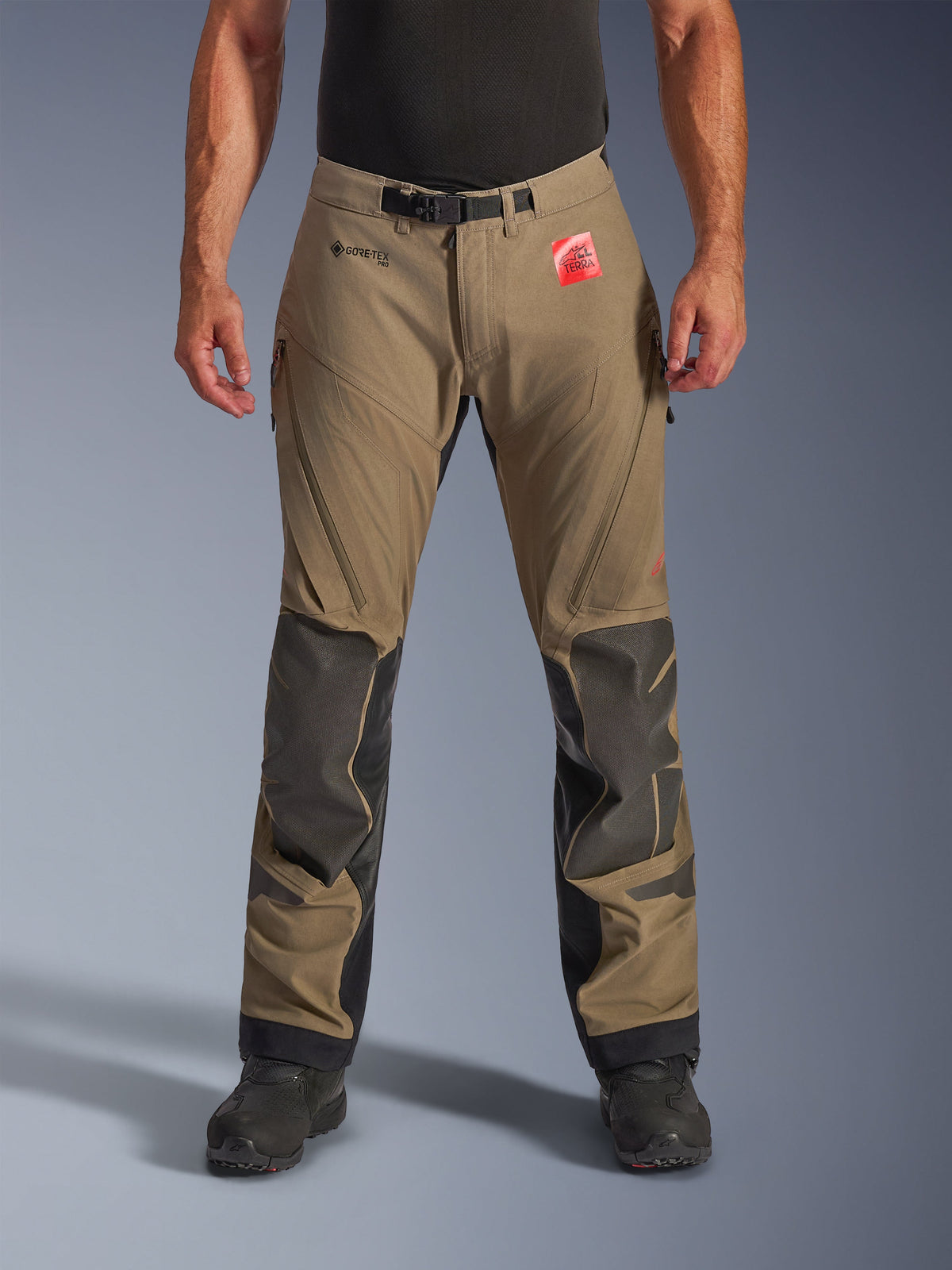 Alpinestars Nazca 3L GORE-TEX PRO broek in Walnut Black, adventure touring motorbroek voor heren, bruin textiel met zwarte Superfabric-kniebeveiliging, ventilatieopeningen met rits op de dijen en magnetische taillesluiting, vooraanzicht op model met zwarte laarzen, duurzame waterdichte motoruitrusting