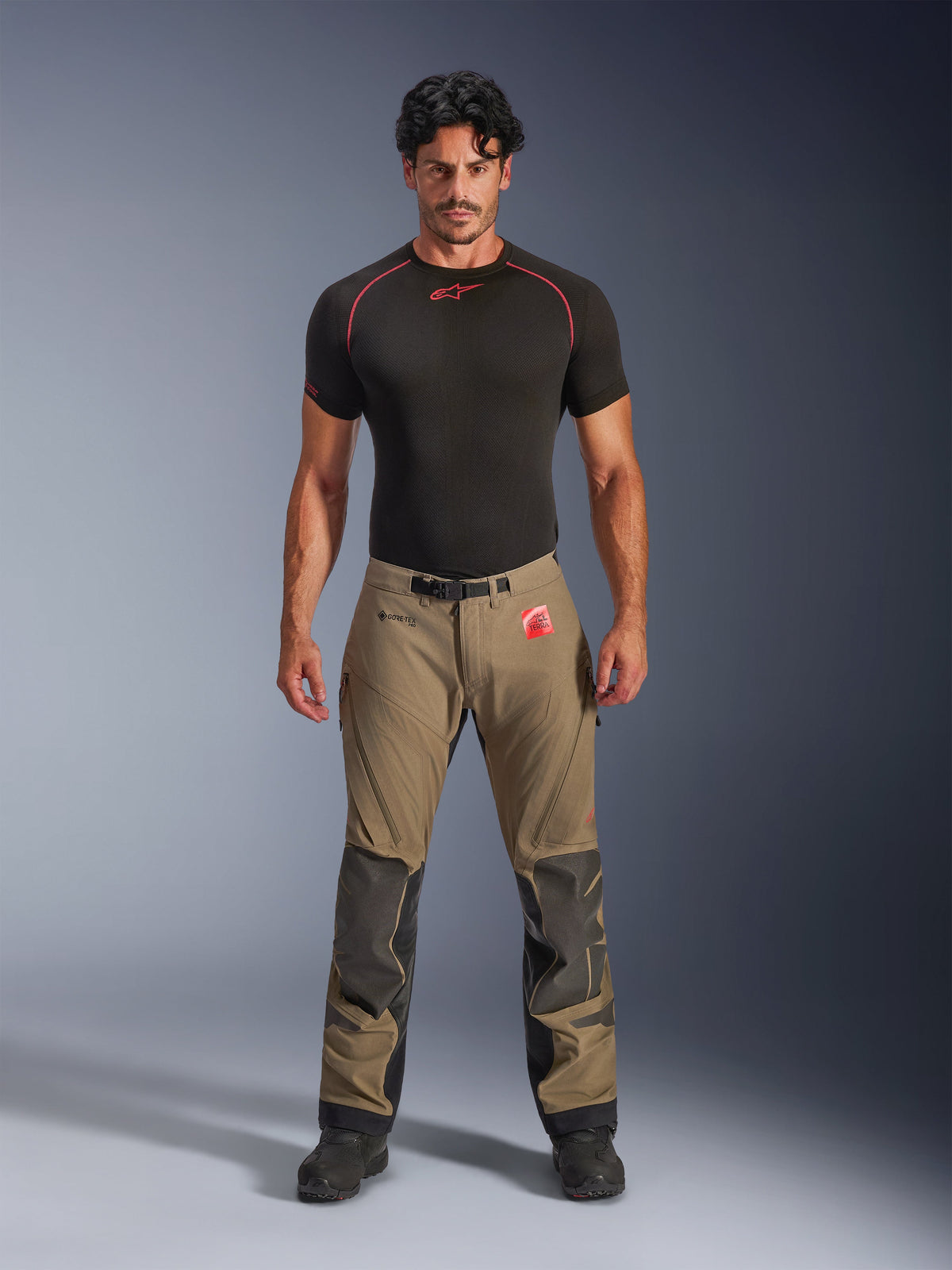 Mannelijk model in Alpinestars Nazca 3L GORE-TEX PRO broek in walnootbruin en zwart, technische adventure-rijbroek met Superfabric knieversterkingen en een magnetische gespsluiting, gecombineerd met een zwart Alpinestars base layer shirt, studio vooraanzicht.