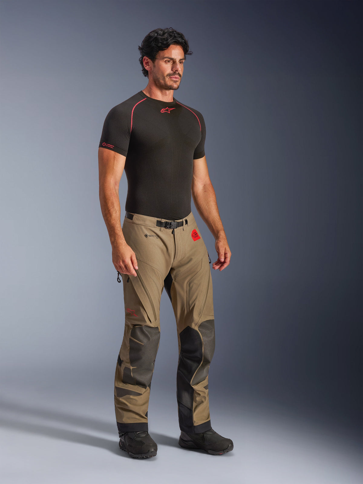 Alpinestars Nazca 3L GORE-TEX PRO broek in Walnut Black, adventure motorbroek met zwarte Superfabric-versterkingen op de knieën, gedragen door een model met een zwarte compressie-onderlaag, voorzien van een magnetische gespriem en rode All Terra-branding.