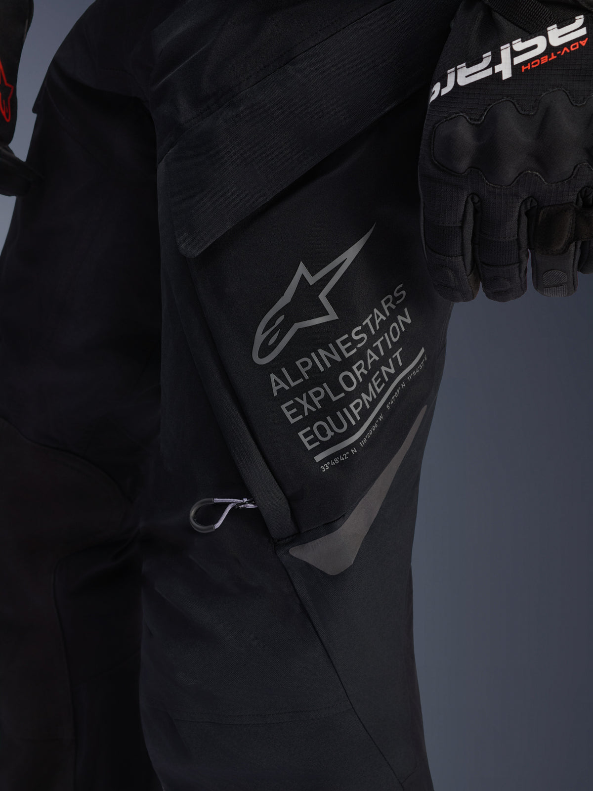 Close-up van de Alpinestars ST-7 2L GORE-TEX broek in het zwart, gedetailleerde weergave van de grote cargozak op de dij met grijze Alpinestars Exploration Equipment branding, sterlogo en geografische coördinaten, hoogwaardige textielconstructie voor adventure touring.