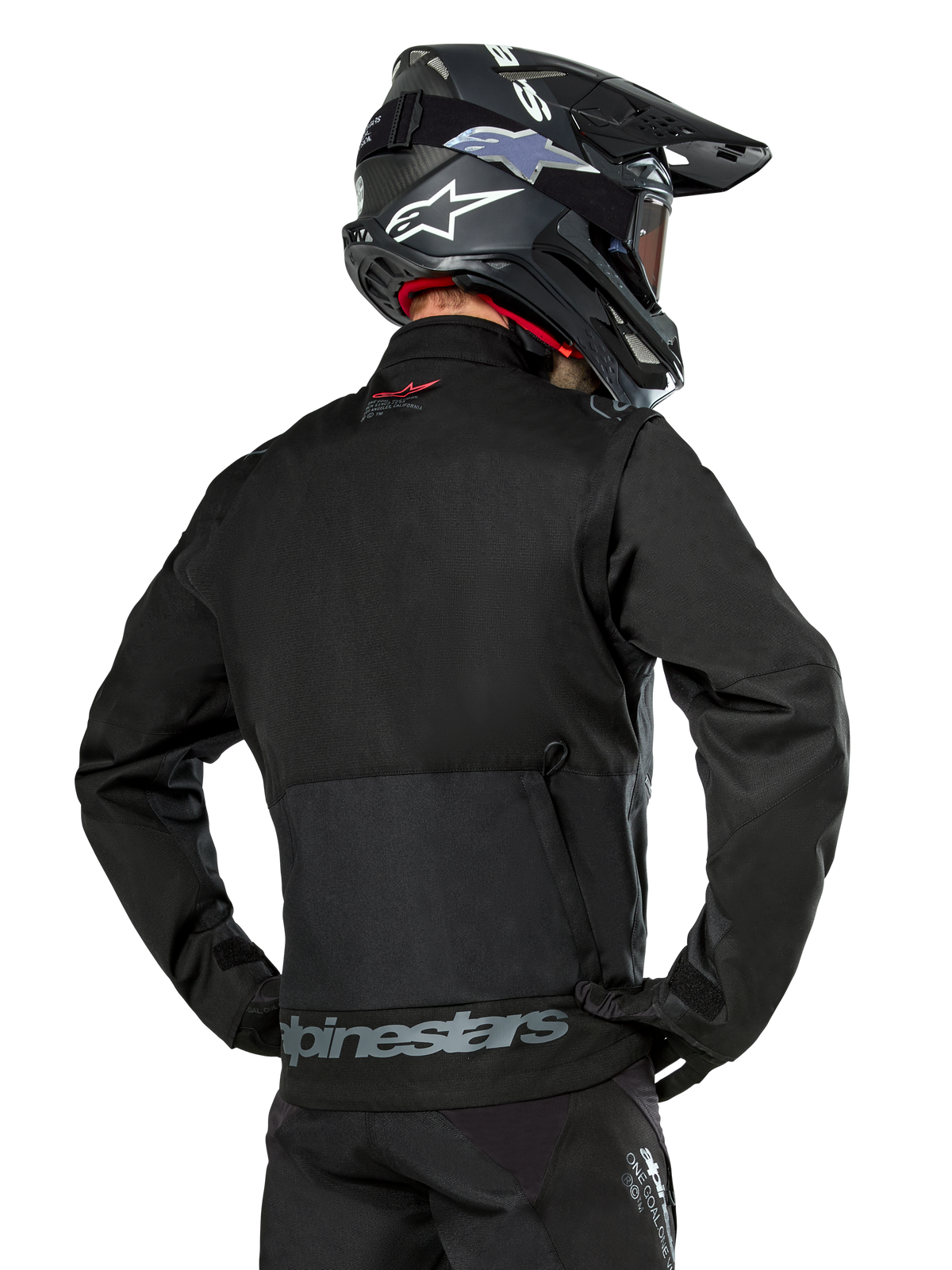 Achteraanzicht van een rijder die de Alpinestars Pro-Dura jas in het zwart draagt, duurzame ripstop textiel enduro-jas, voorzien van een grote cargo-zak met grijze Alpinestars-branding op de onderrug, gecombineerd met een zwarte motorcrosshelm.