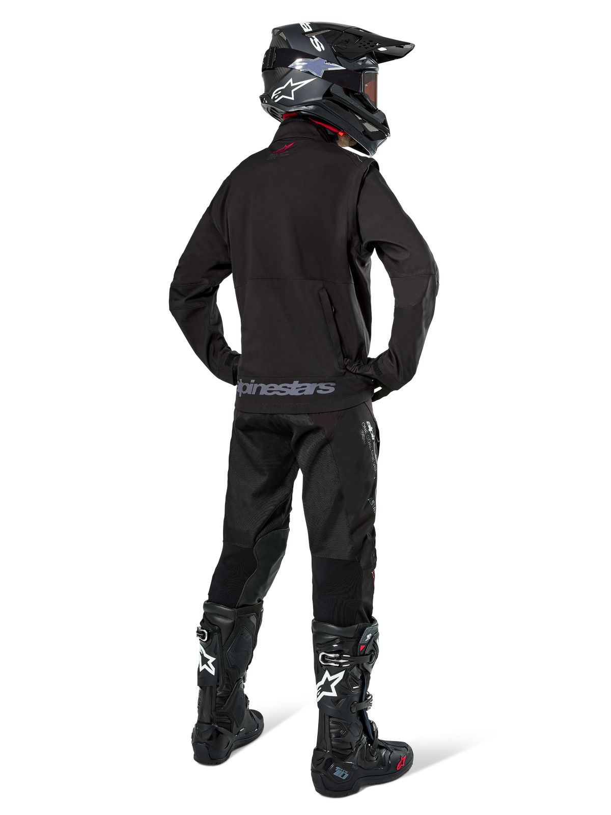Enduro rider viewed from the back wearing the Alpinestars Lite-Dura Softshell Jacket in Black, paired with black technical riding pants and boots, featuring a zippered rear storage pocket and Alpinestars logo on the lower hem, complete off-road gear for motocross.