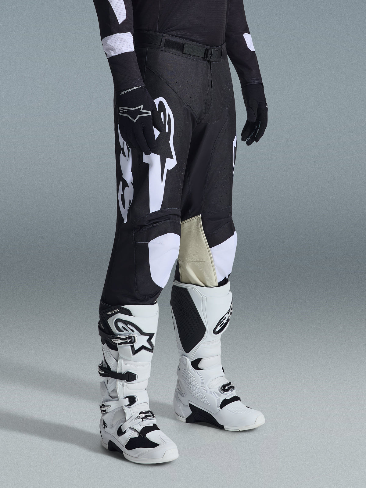 Alpinestars 2026 Racer Air Riway broek, motorcrossbroek, zwart en wit, gedragen met witte en zwarte Tech Series laarzen en een zwart shirt met lange mouwen, voorzien van witte Alpinestars-branding, lasergesneden perforaties en beige geitenleren binnenknieën voor off-road prestaties.