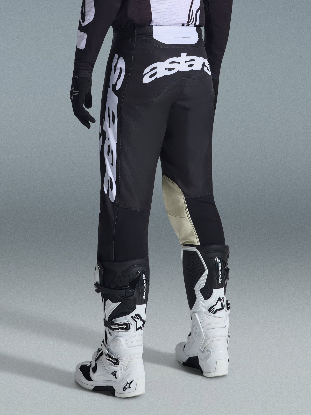 Alpinestars 2026 Racer Air Riway Pants, motorcrossbroek, zwart en wit, achteraanzicht met grote witte Astars-merknaam op het zitvlak en de benen, beige lederen panelen aan de binnenkant van de knieën, gedragen met witte en zwarte Tech 7 laarzen, hoogwaardige off-road racekleding.