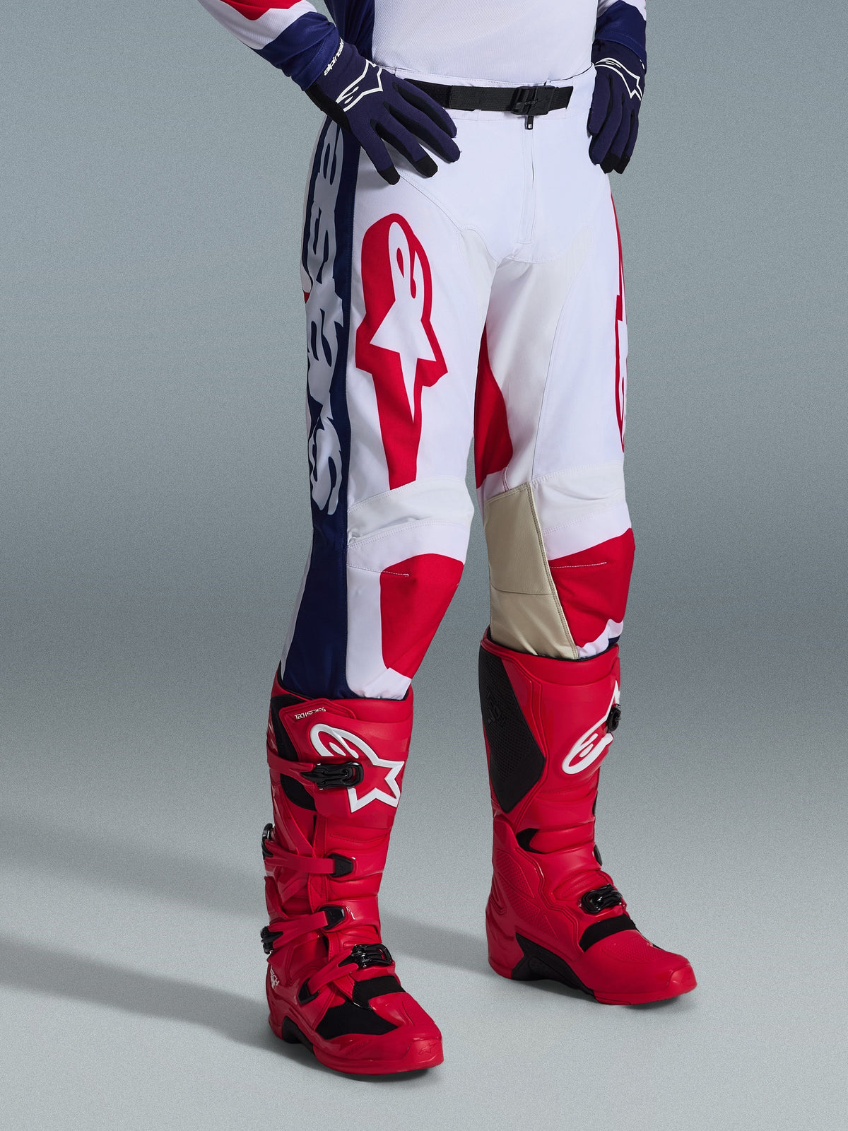 Alpinestars 2026 Racer Riway broek, motorcrossbroek, wit blauw rood, met opvallende rode logo's op de dijen en blauwe zijpanelen met witte merknaam, gedragen met rode laarzen en donkerblauwe handschoenen, ergonomisch ontwerp voor off-road racen