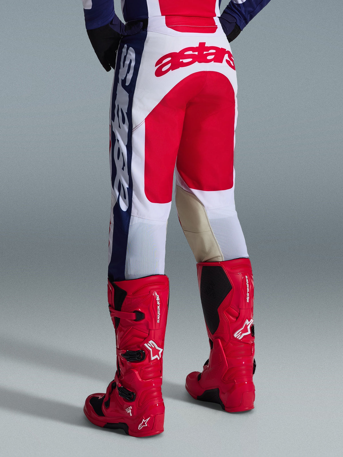 Alpinestars 2026 Racer Riway crossbroek, motorcrossbroek, wit, blauw en rood, achteraanzicht met groot rood Astars-logo op het zitvlak en beige lederen panelen aan de binnenzijde van de knieën, gedragen met rode Alpinestars motorcrosslaarzen, technische kleding voor off-road racen