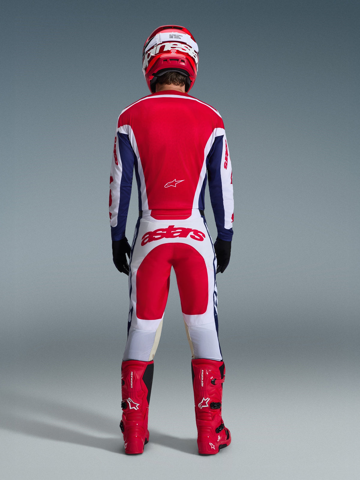 Motorcrosser van achteren gezien in volledige race-uitrusting, Alpinestars 2026 Racer Riway-broek in wit, blauw en rood met bijpassende jersey, rode laarzen en helm, voorzien van een groot 'astars'-logo op het zitvlak, gecoördineerde motorcross-outfit.
