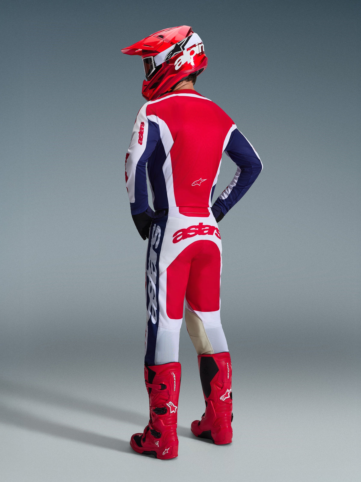 Motorcrosser van achteren gezien die de Alpinestars 2026 Racer Riway Pants draagt in wit, blauw en rood, gecombineerd met een bijpassende jersey, rode helm en felrode laarzen, waarbij de opvallende branding op het zitvlak en de atletische off-road pasvorm zichtbaar zijn.