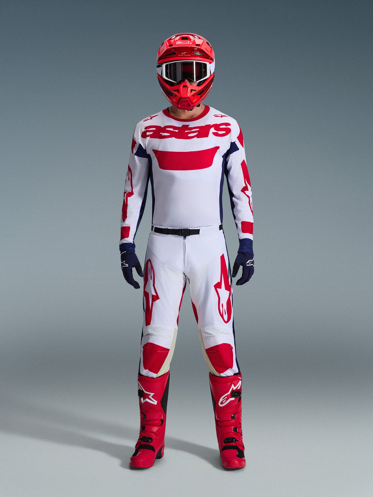 Motocross-rijder in volledige race-uitrusting, wit jersey met rode Alpinestars-branding en blauwe accenten, Alpinestars 2026 Racer Riway broek in White Blue Red, rode helm met bijpassende crossbril, rode en zwarte laarzen, marineblauwe handschoenen, gecoördineerde outfit voor off-road racen