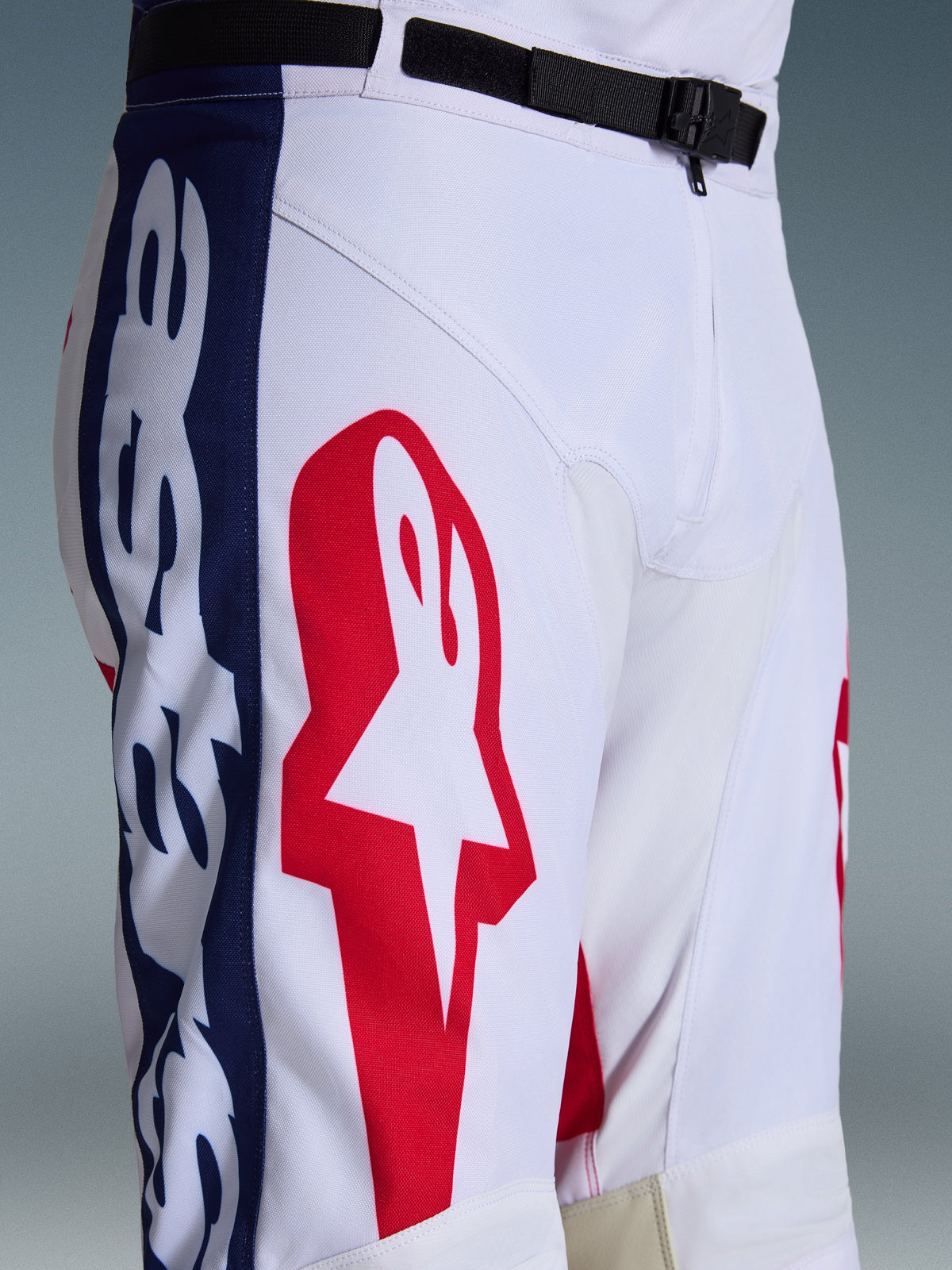 Alpinestars 2026 Racer Riway broek, motorcrossbroek, wit blauw rood, close-up van het bovenbeen en de taille met groot rood sterlogo op wit polyfabric, blauwe zijpanelen met branding en zwarte verstelbare tailleband met SX2 magnetische gesp, sportief off-road racing ontwerp