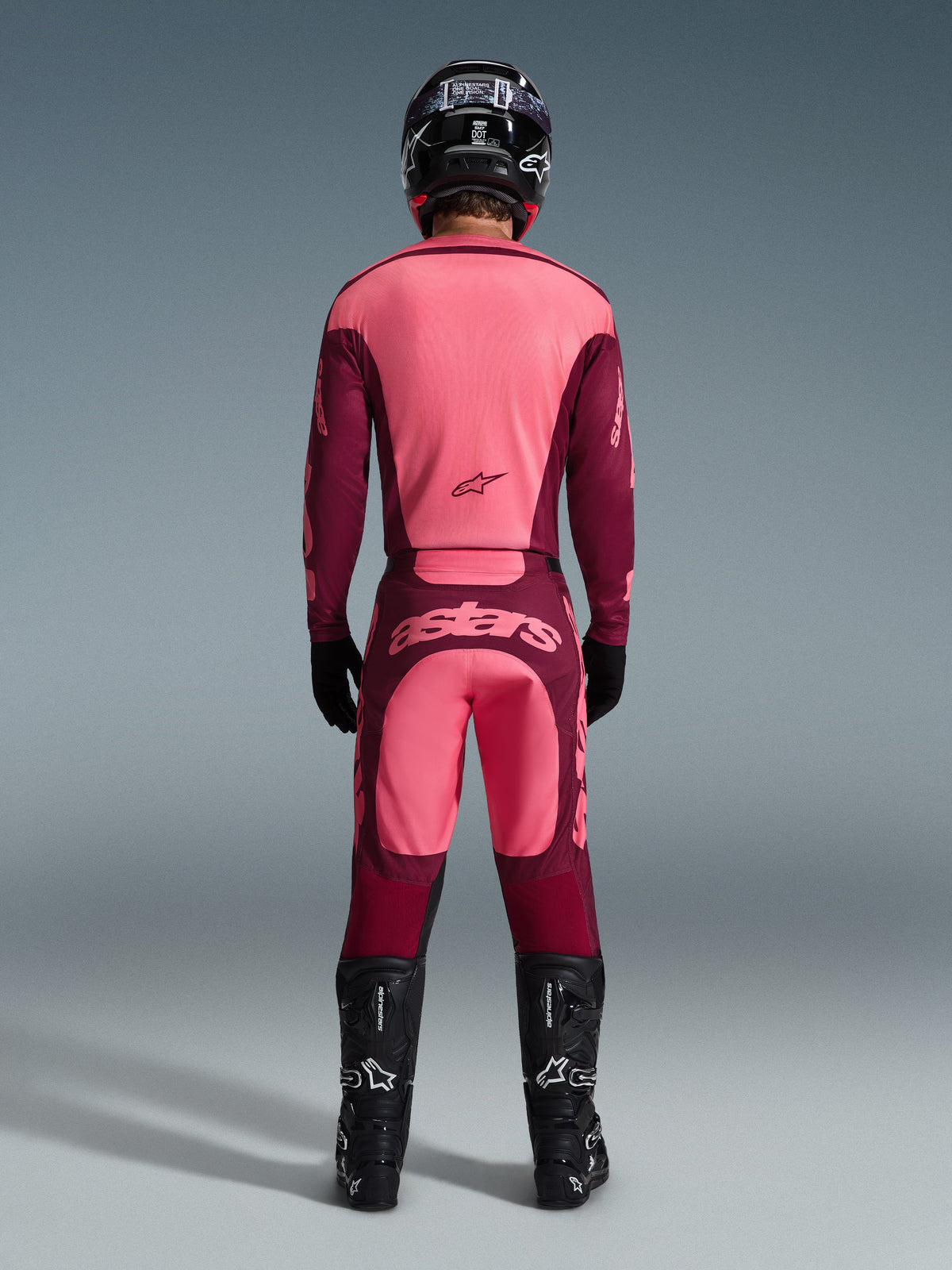 Rear view of a motocross rider wearing the 2026 Racer Riway Gear Set, pink and burgundy jersey, matching burgundy pants with light pink 'astars' logo on the seat, black helmet, black boots, and black gloves, coordinated outfit for off-road performance