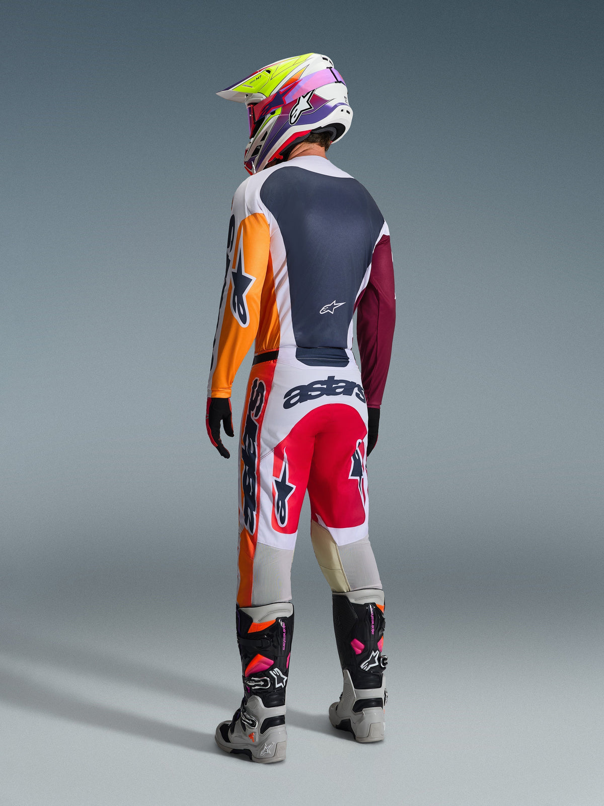 Motorcrosser van achteren gezien in volledige race-uitrusting, Alpinestars 2026 Racer Portl-broek in Red Light Gray en Dark Gray, bijpassende jersey met oranje en kastanjebruine mouwen, meerkleurige grafische helm, grijze en zwarte off-road laarzen met roze accenten, professionele motorcrossoutfit