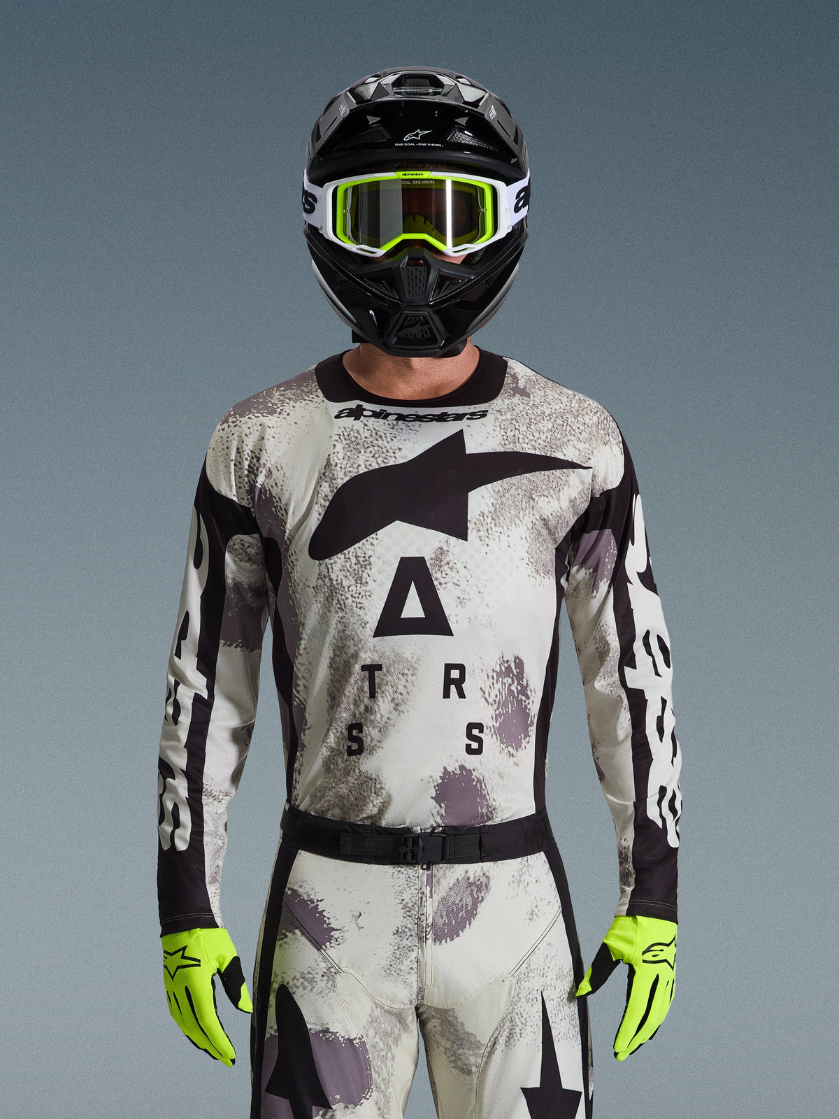 Alpinestars 2026 Racer Lahnd Jersey, motorcross kledingset, Sand Camo-patroon in grijs en wit, groot zwart sterlogo en 'ASTARS'-belettering, gedragen met zwarte helm, neongele crossbril en neongele handschoenen, professionele off-road race-uitrusting