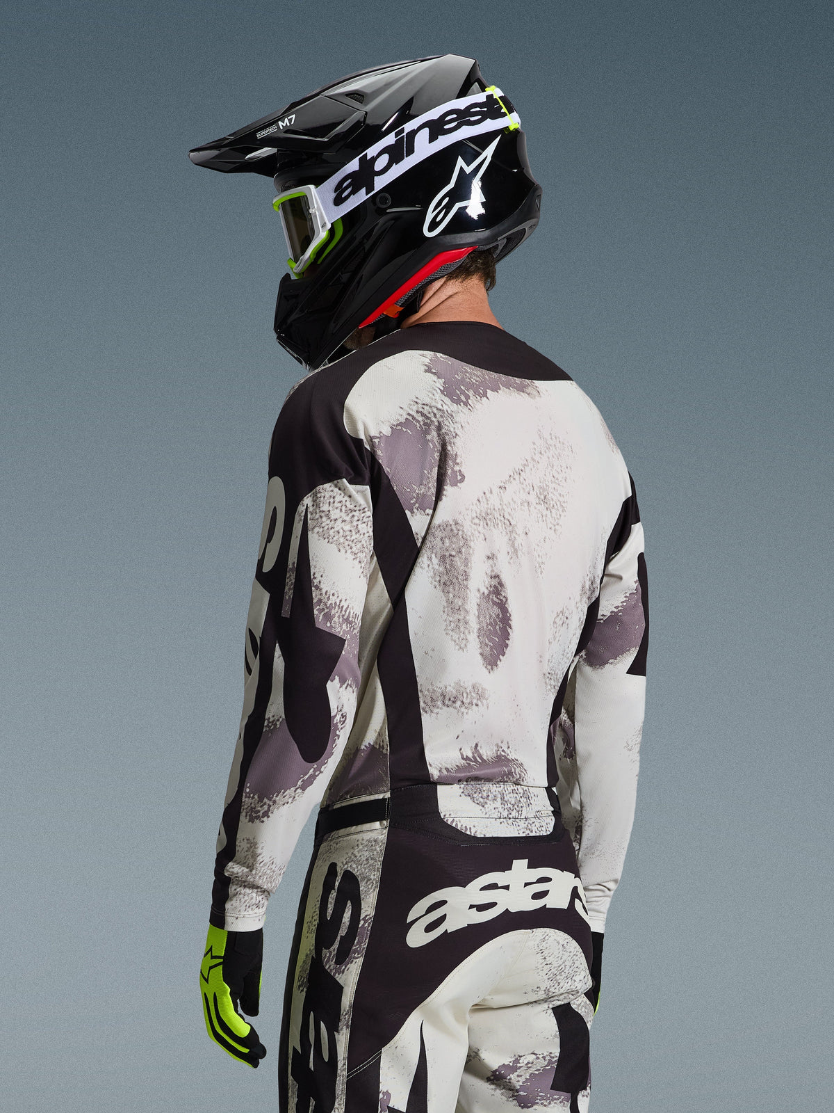 Motorcrosser van achteren gezien in de Alpinestars 2026 Racer Lahnd Gear Set, grijs en wit Sand Camo jersey en broek met zwarte accenten, zwarte helm met wit Alpinestars-logo, neongele en zwarte handschoenen, gecoördineerde off-road performance outfit
