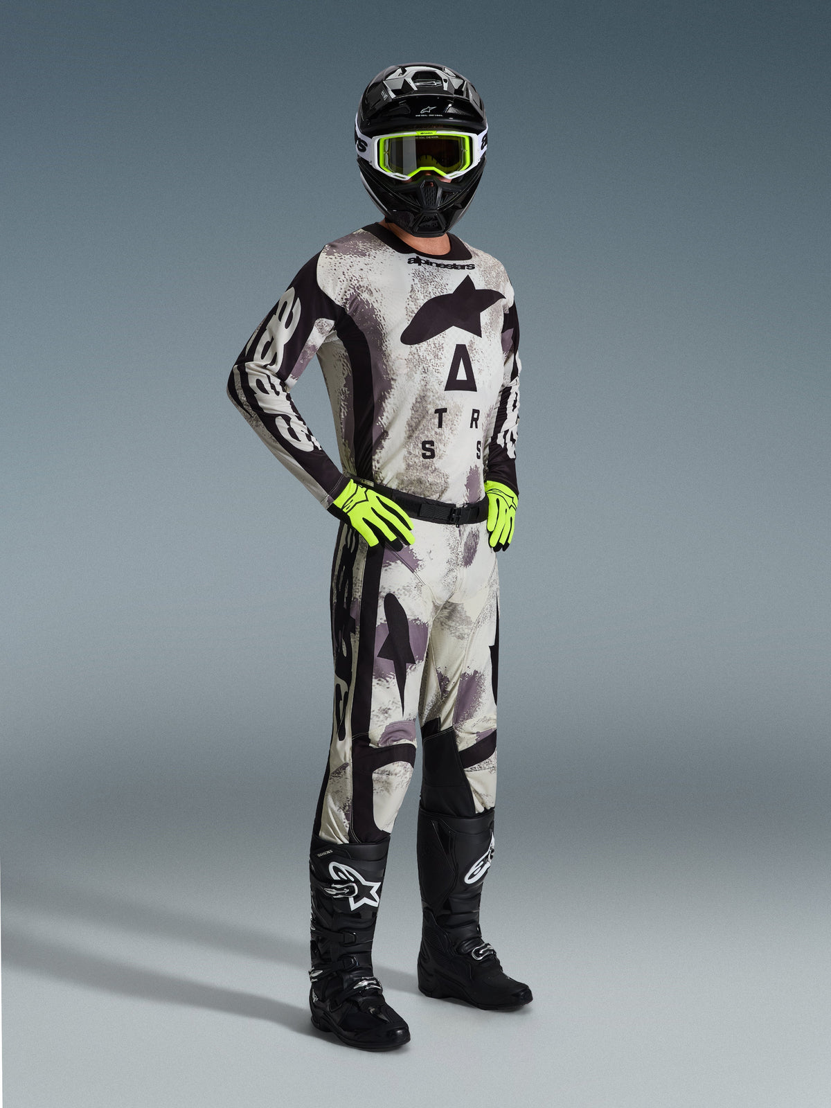 Alpinestars 2026 Racer Lahnd kledingset, motorcrossrijder in volledige race-uitrusting in Sand Camo en grijs, bijpassende jersey en broek met zwarte accenten en opvallende branding, neongele crossbril en handschoenen, zwarte helm en zwarte laarzen.
