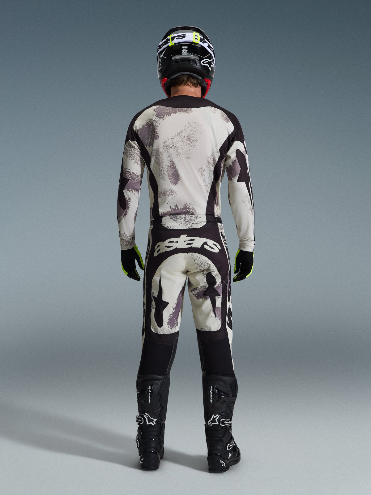 Motorcrossrijder in complete Alpinestars 2026 Racer Lahnd gearset, van achteren gezien, Sand Camo jersey en broek met lichtgrijs en zwart abstract patroon, zwarte helm, zwarte en neongele handschoenen, zwarte laarzen, voorzien van 'astars' branding op de achterzijde van de broek