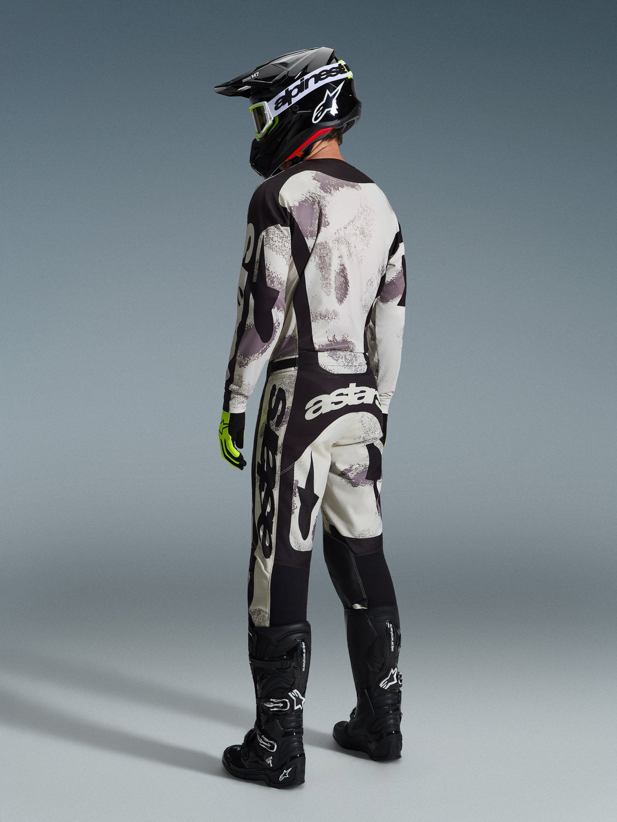 Motocrossrijder in Alpinestars 2026 Racer Lahnd Gear Set, Sand Camo abstract patroon in grijs en wit, zwarte broek met opvallende witte Alpinestars-branding, zwarte helm en laarzen, fluorescerende groene accenten op de handschoenen, complete outfit voor off-road racing
