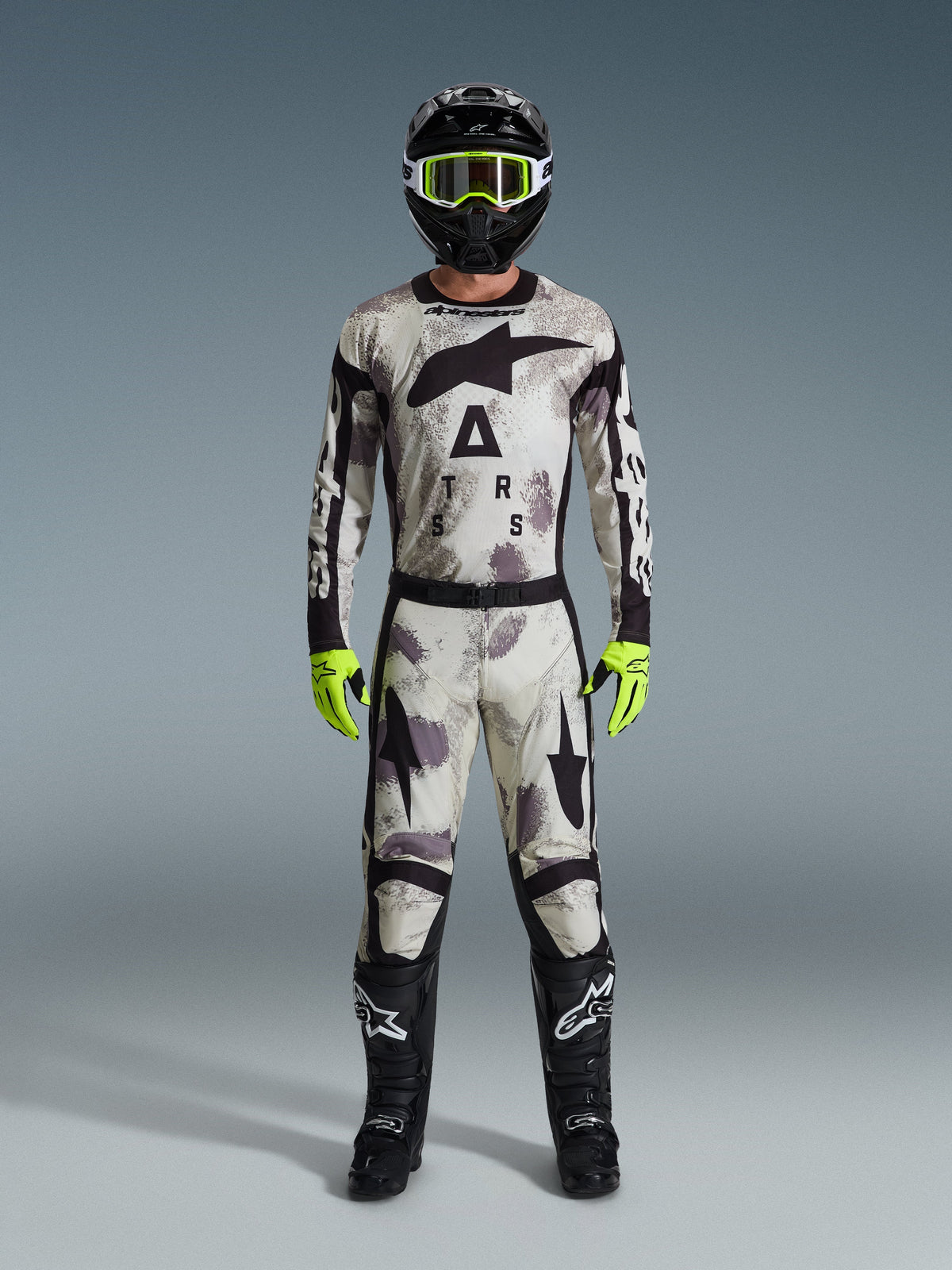 Motocrossrijder in de Alpinestars 2026 Racer Lahnd Gear Set, pak met Sand Camo-patroon in wit, grijs en zwart, zwarte helm met neongroene crossbril, fel neongroene handschoenen en zwarte laarzen met witte logo's, complete gecoördineerde outfit voor off-road racing