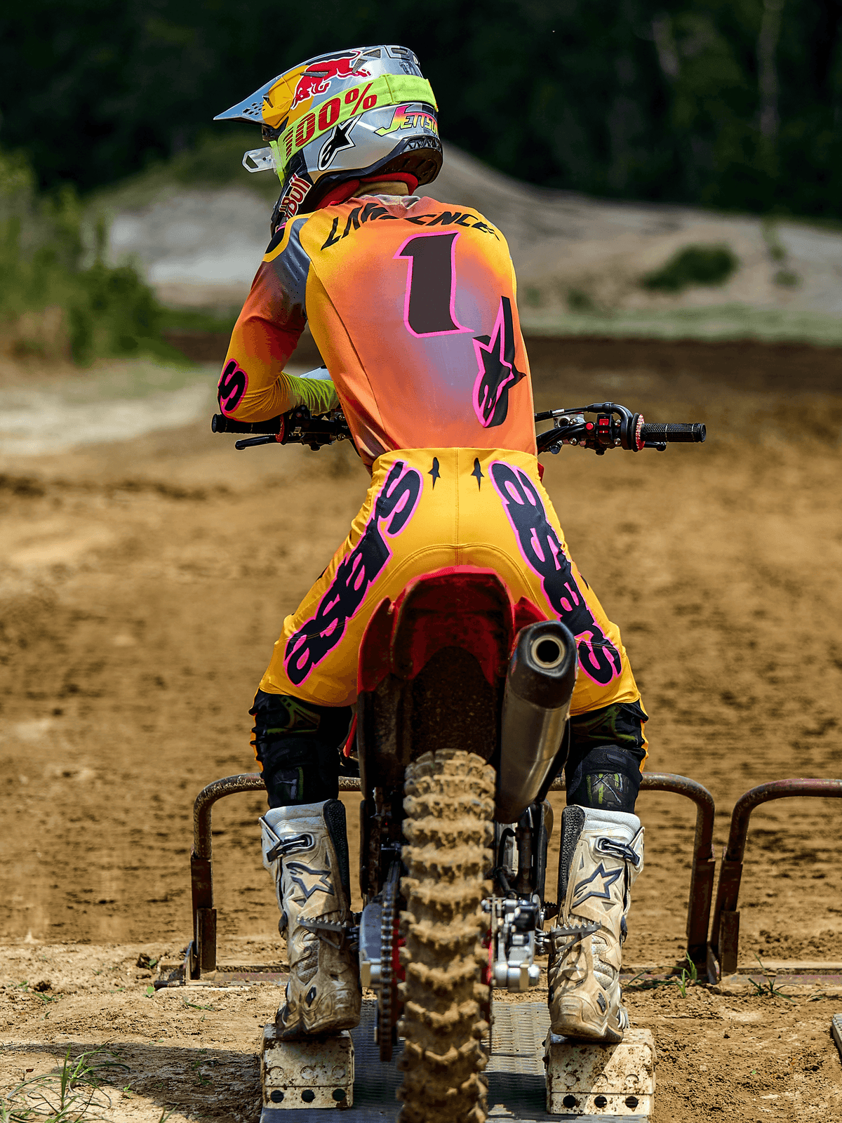 Motorcrosser van achteren in Alpinestars 2025 Supertech Lipan Gear Set, geel en roze, jersey met nummer 1 en 'LAWRENCE' op de rug, gele broek met opvallende roze 'ASTARS' branding, witte laarzen en bijpassende helm, zittend op een crossmotor bij het starthek