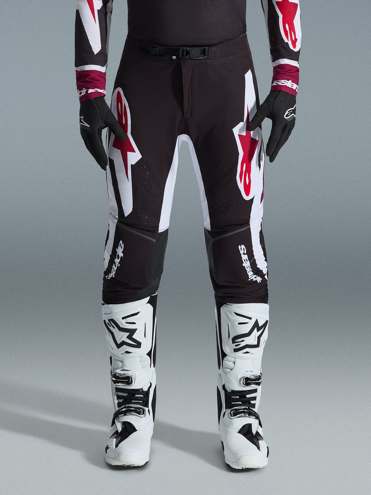 Alpinestars 2026 Supertech AFD Vista Broek, motorcrossbroek, zwart, rood en wit, met grote grafische logo's op de bovenbenen en knieën, gedragen met witte racelaarzen en zwarte handschoenen, atletisch race-ready ontwerp voor elite off-road competitie