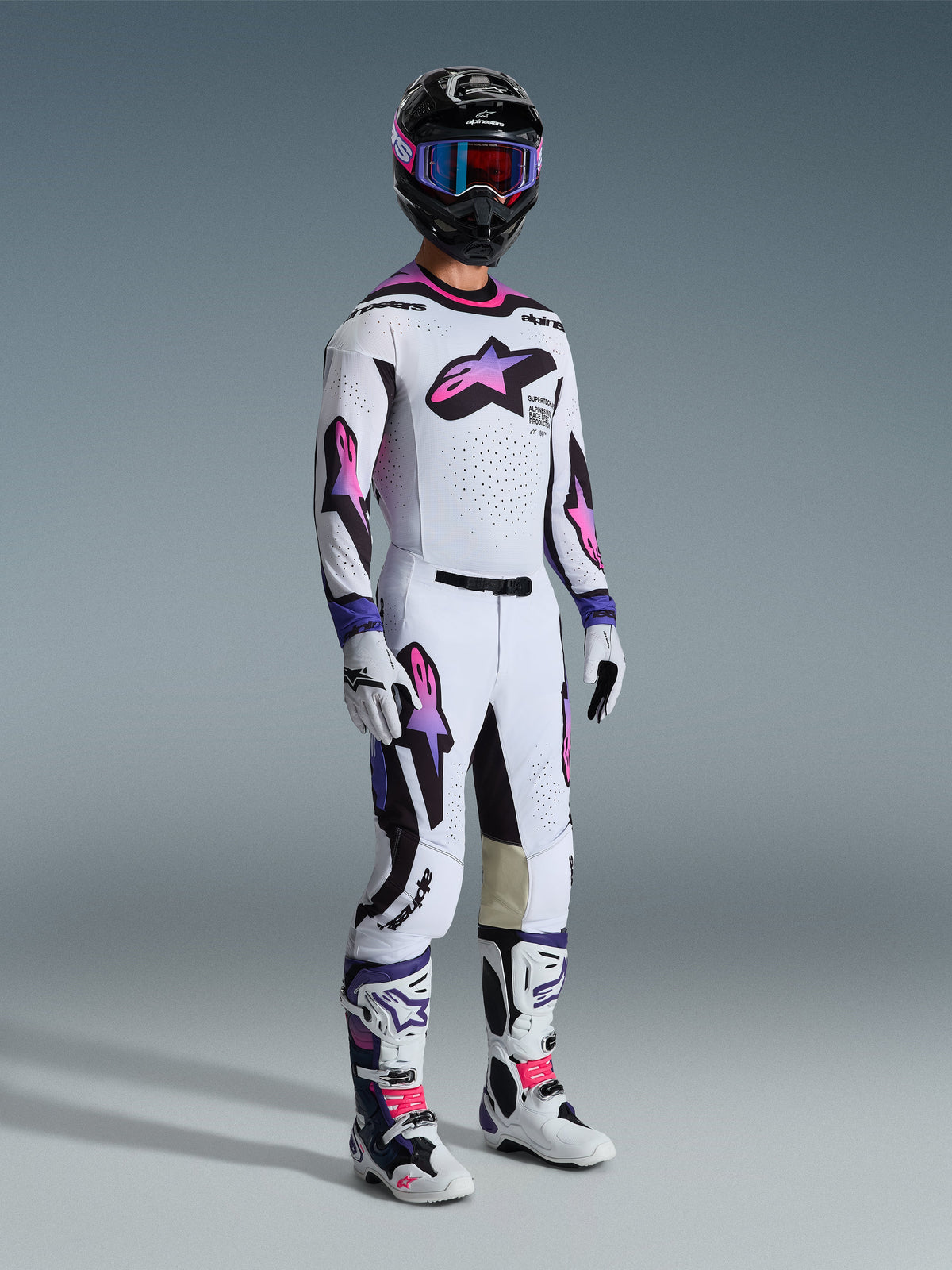 Motorcrossrijder in een complete Alpinestars race-uitrusting, met de 2026 Supertech AFD Vista-broek in wit, paars en zwart, bijpassende jersey, zwarte helm met paarse bril, witte en paarse laarzen en witte handschoenen, gecoördineerde professionele off-road race-uitrusting.