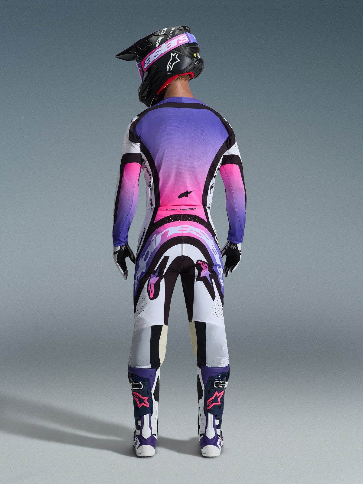 Motocrossrijder van achteren gezien in volledige race-uitrusting, shirt met paars en roze kleurverloop, Alpinestars 2026 Supertech AFD Vista broek in White Purple Black, zwarte helm met paarse crossbril, witte en paarse laarzen, gecoördineerde off-road racekit.