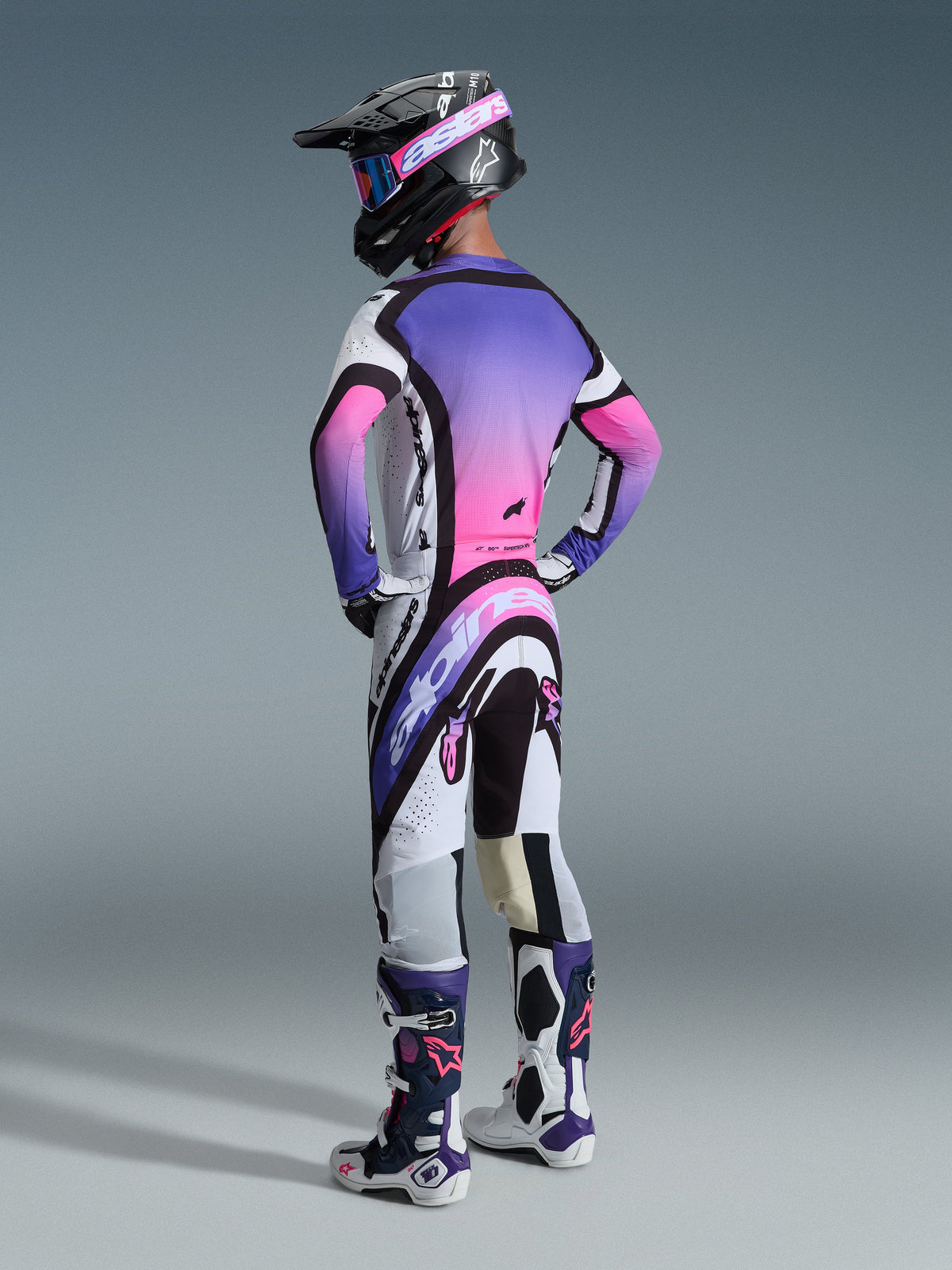 Motocrossrijder van achteren gezien in een Alpinestars Supertech AFD Vista broek 2026, kleurstelling wit, paars en zwart, gecombineerd met een bijpassende paars-roze gradient jersey, zwarte helm en wit-paarse racelaarzen voor een complete off-road racing look.