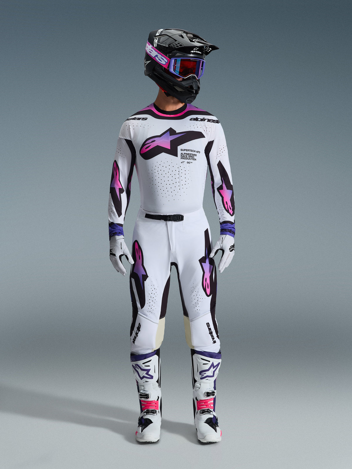 Motorcrossrijder met een complete race-uitrusting, Alpinestars 2026 Supertech AFD Vista broek en jersey in wit, paars en zwart, bijpassende handschoenen, witte en zwarte laarzen, zwarte helm met paarse bril, strakke atletische pasvorm voor off-road prestaties