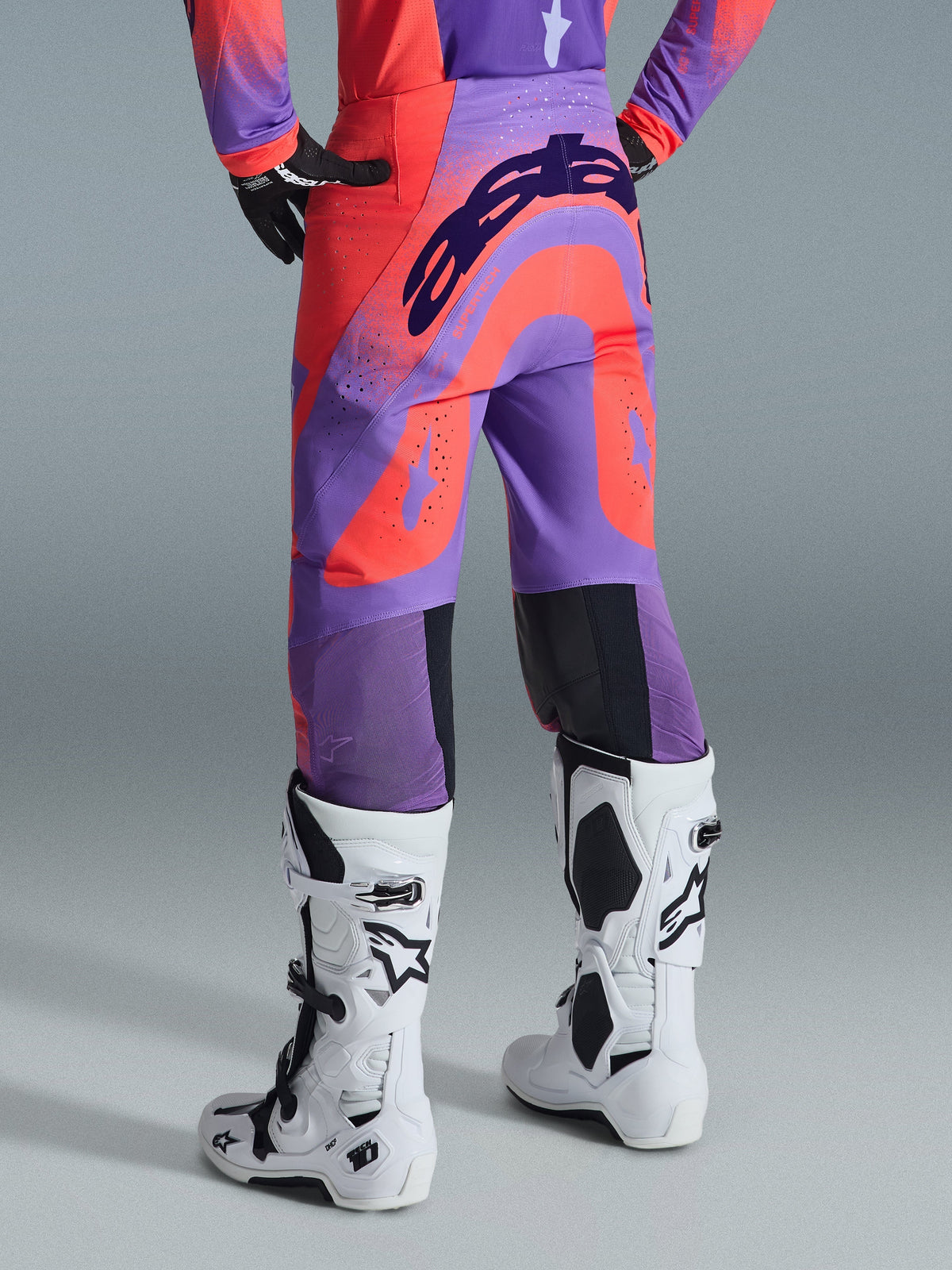 Alpinestars 2026 Supertech Scenz Pants, Motocross Pants, Hot Coral Purple, rear view with bold branding across the seat, vibrant coral and purple panels with perforated zones for airflow, worn with white and black racing boots