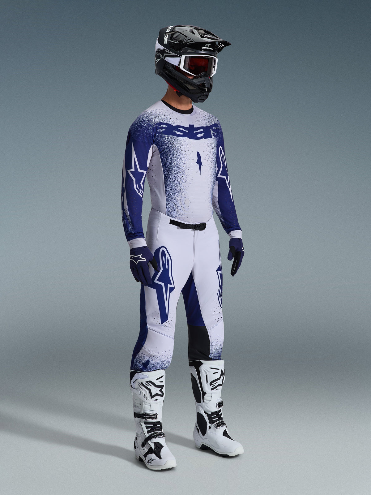 Alpinestars motorcrossrijder in 2026 Supertech Scenz broek, lichtgrijs en donkerblauw, bijpassende blauw-witte jersey met 'astars' branding en spray-paint effect, zwarte helm met witte goggle en witte en zwarte motorcrosslaarzen, gecoördineerde professionele off-road race-outfit.