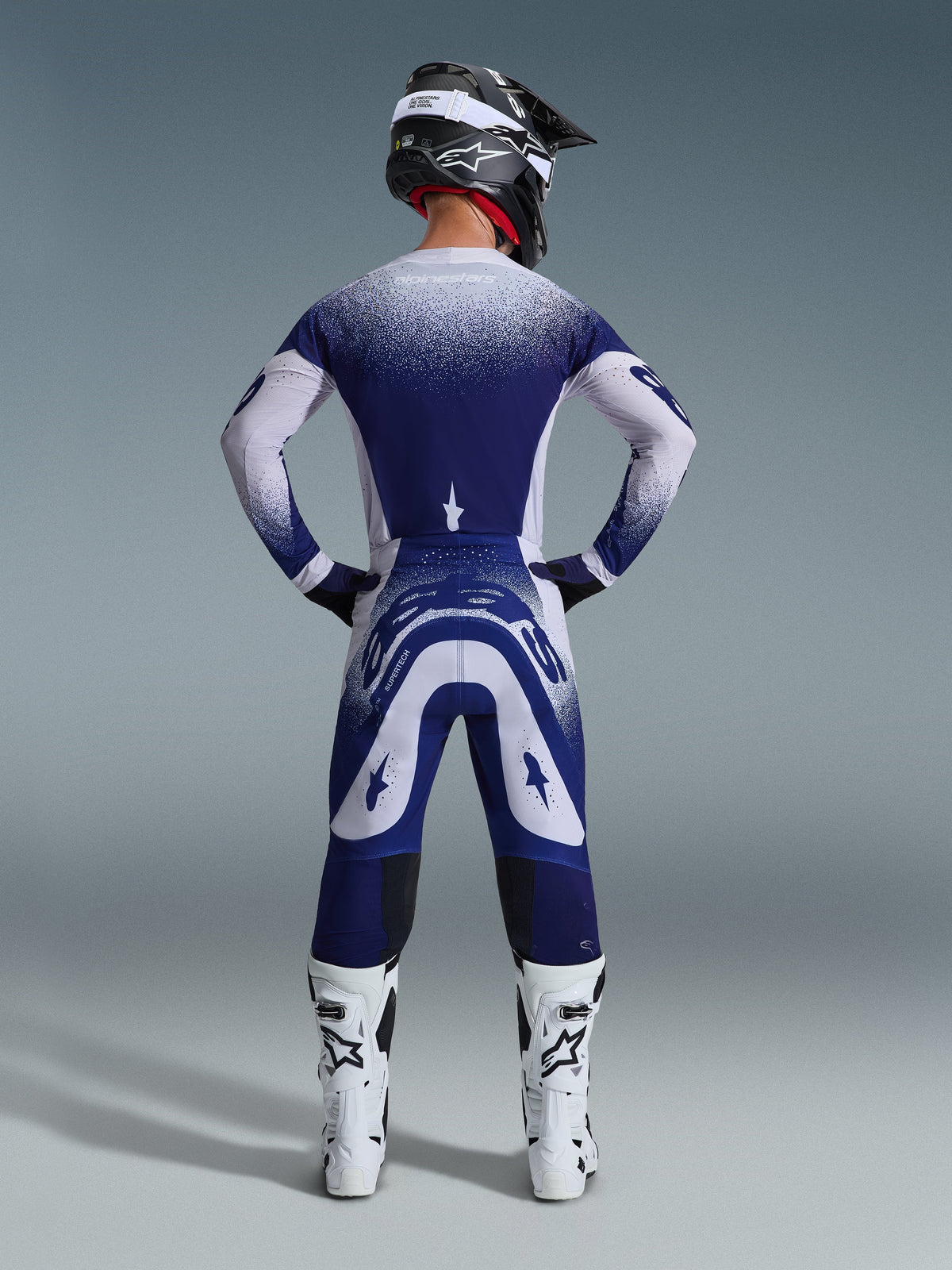 Alpinestars 2026 Supertech Scenz broek, motorcrossbroek, lichtgrijs en donkerblauw, achteraanzicht van een rijder in een bijpassende kledingset met gespikkelde kleurverloop-graphics, witte laarzen en zwarte helm, professionele atletische pasvorm voor off-road racing