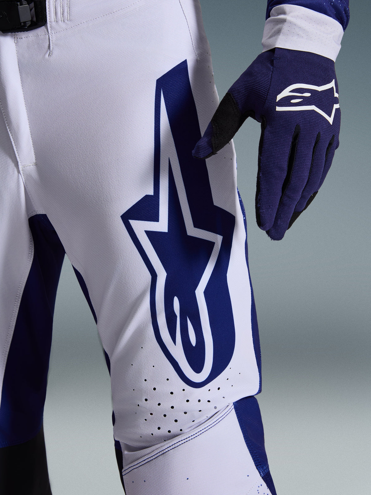 Alpinestars 2026 Supertech Scenz broek, motorcrossbroek, lichtgrijs en donkerblauw, close-up van de dij met een groot donkerblauw Alpinestars-logo, geperforeerde ventilatiezones en stretch rip-stop materiaal, gedragen met een bijpassende donkerblauwe motorcrosshandschoen