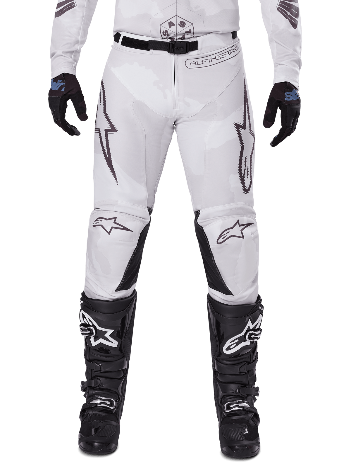 Alpinestars 2025 Racer Hollow crossbroek, motorcrossbroek, licht camo bruin, gedragen met zwarte handschoenen en motorcrosslaarzen, voorzien van een subtiel camouflagepatroon met zwarte grafische prints en Astars-logo's, ergonomisch ontwerp voor off-road rijprestaties