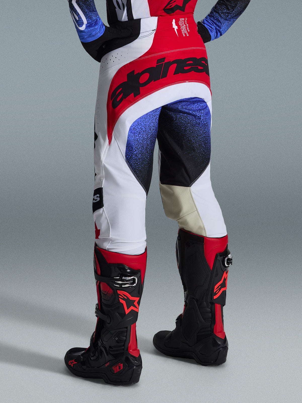Alpinestars 2026 Techstar Knif broek, motorcross broek, wit, rood en blauw, achteraanzicht met rood zitvlak en zwarte branding, blauwe en zwarte gespikkelde patronen op de pijpen, gedragen met zwarte en rode Tech 10 laarzen, elite off-road race-uitrusting
