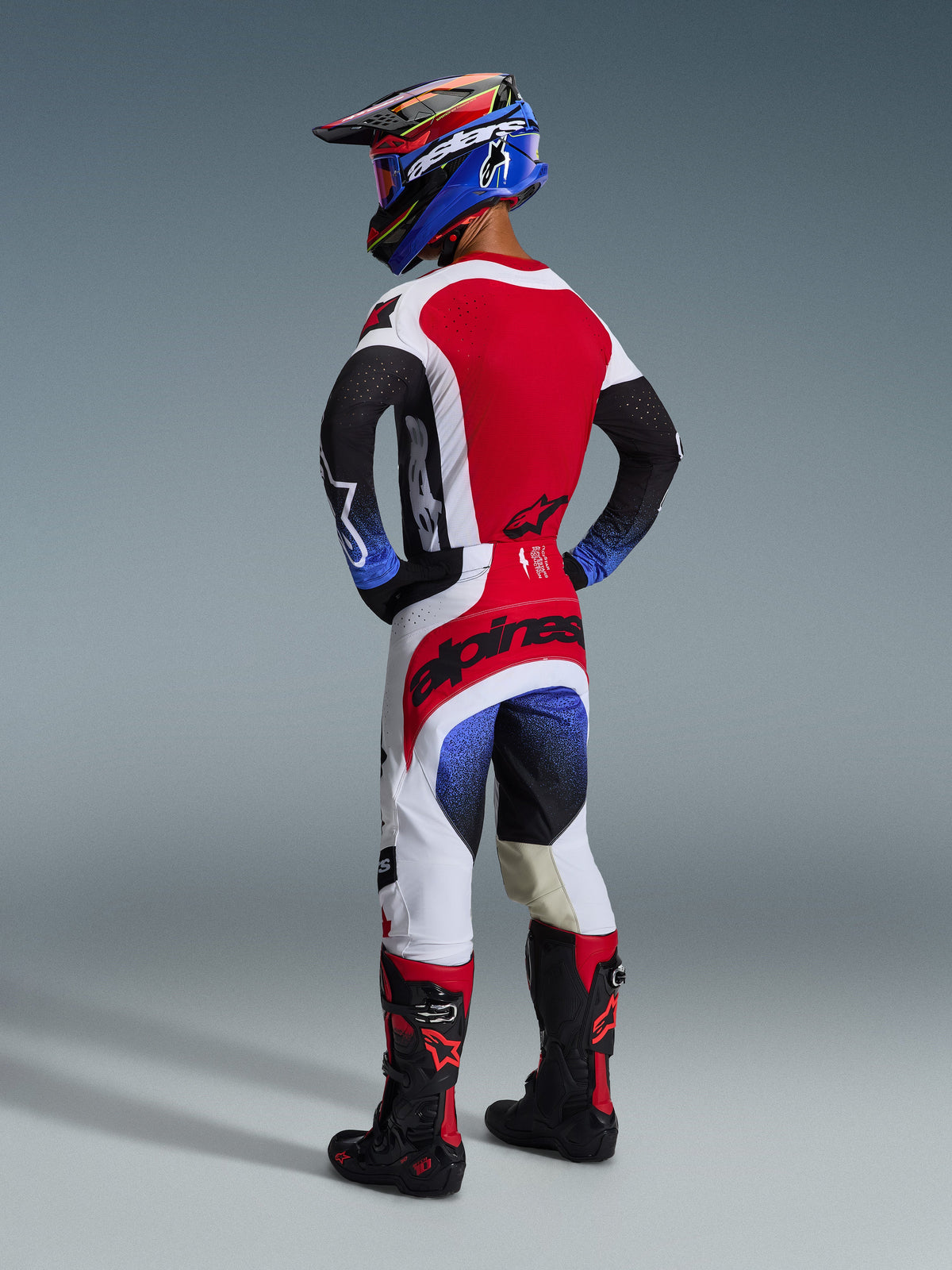 Alpinestars 2026 Techstar Knif broek, motorcrossbroek, zwart, wit en rood met blauwe kleurverloopaccenten, achteraanzicht van rijder in volledige race-uitrusting, opvallende rode branding op het zitvlak, witte en zwarte panelen, gedragen met bijpassende jersey en Tech 10 laarzen