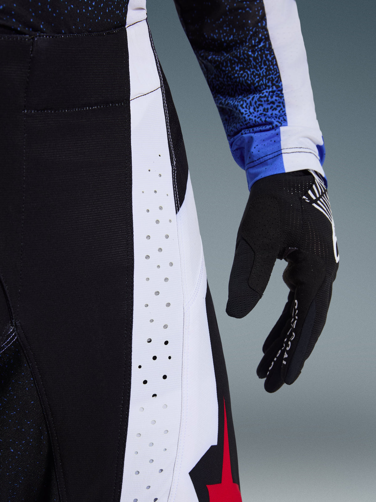 Detailopname van de Alpinestars 2026 Techstar Knif broek in zwart, wit en rood, met lasergesneden perforaties op een wit zijpaneel voor ventilatie, zwarte motorcrosshandschoen en blauwe jersey mouw met patroon, hoogwaardige raceconstructie