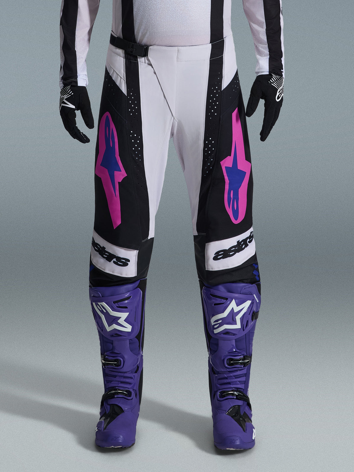 Motocrossrijder in de Alpinestars 2026 Techstar Knif Gear Set, wit-zwarte broek met roze en blauwe logo's, paarse laarzen met witte branding, zwarte handschoenen, met een atletische racepasvorm en lasergesneden ventilatie voor off-road racing