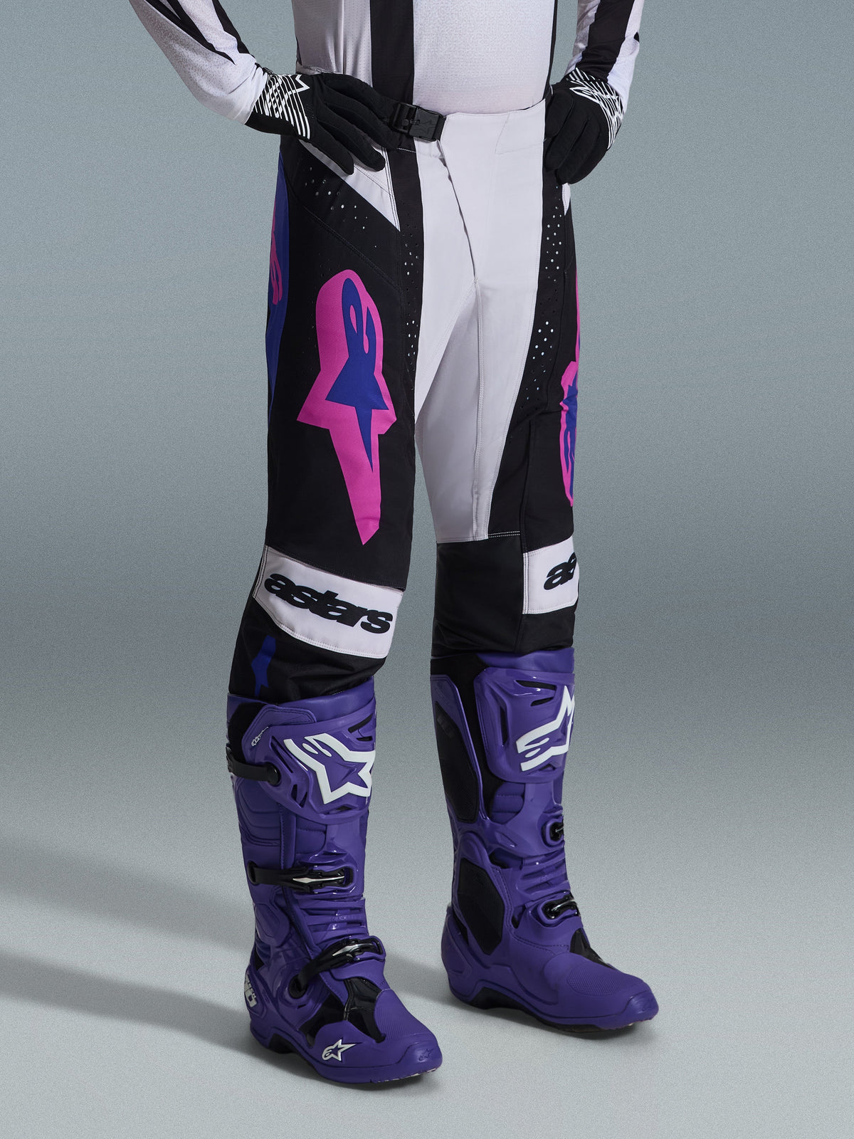 Alpinestars 2026 Techstar Knif broek, zwart, lichtgrijs en paars, gedragen met bijpassende paarse motocrosslaarzen, met opvallende roze accenten en 'astars'-branding, professionele motorcrossuitrusting