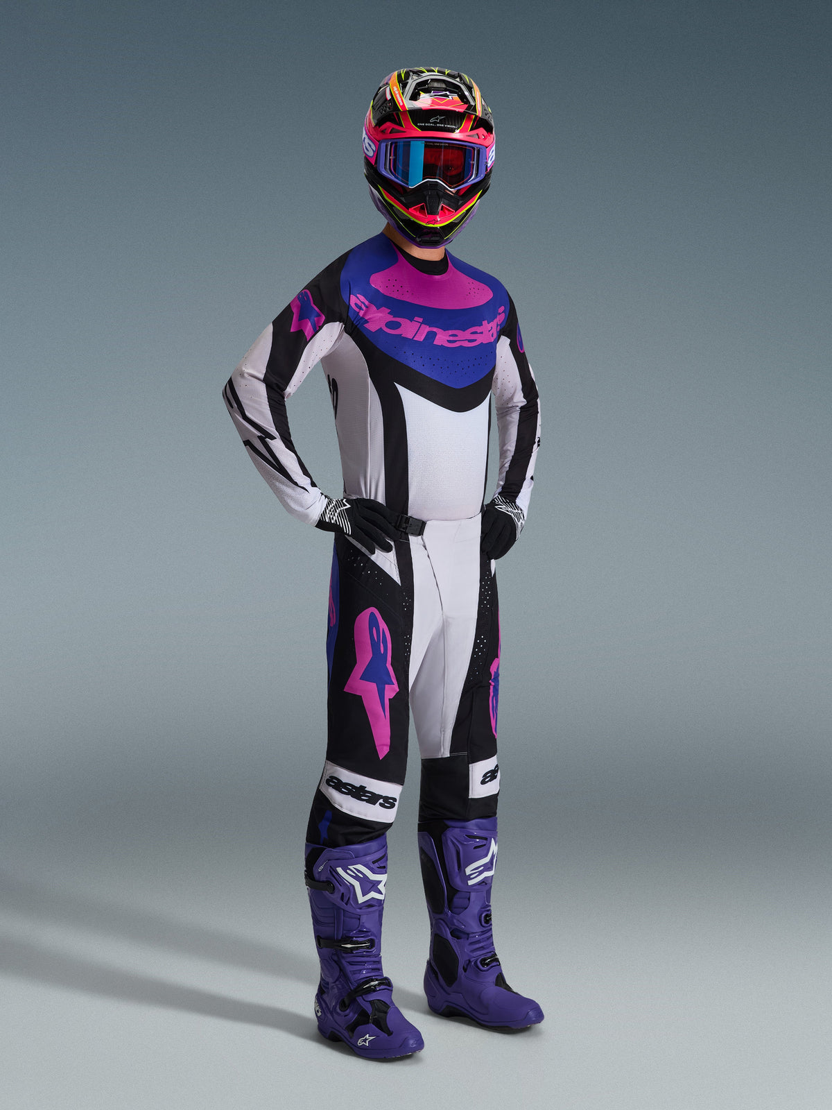 Motocrossrijder in de Alpinestars 2026 Techstar Knif Gear Set, wit jersey met zwarte, blauwe en roze accenten, bijpassende broek met roze Astars-logo's, veelkleurige helm met crossbril, paarse crosslaarzen, gecoördineerde high-performance uitrusting voor motocrosswedstrijden