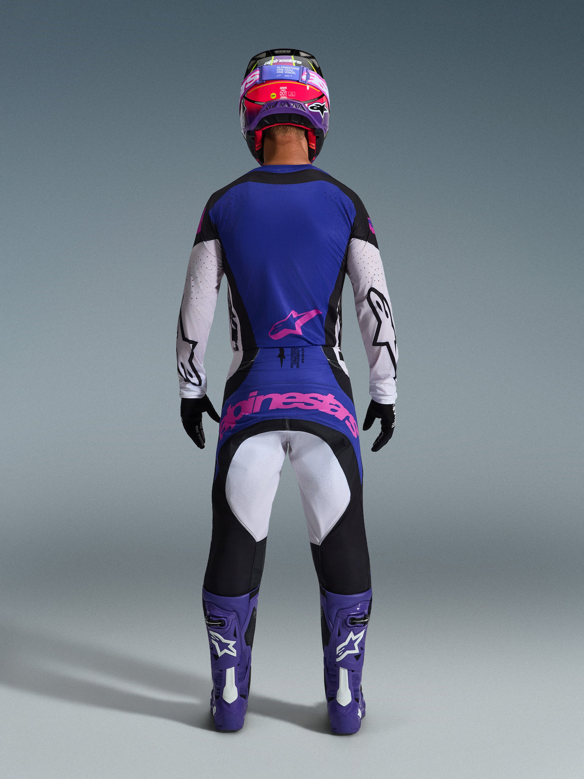 Achteraanzicht van een motorcrossrijder met de Alpinestars 2026 Techstar Knif Gear Set, blauwe en witte jersey met roze ster-logo, bijpassende blauwe en zwarte broek met grote roze Alpinestars-branding op de achterzijde, paarse en witte laarzen en een paarse en rode helm, gecoördineerde professionele off-road race-outfit