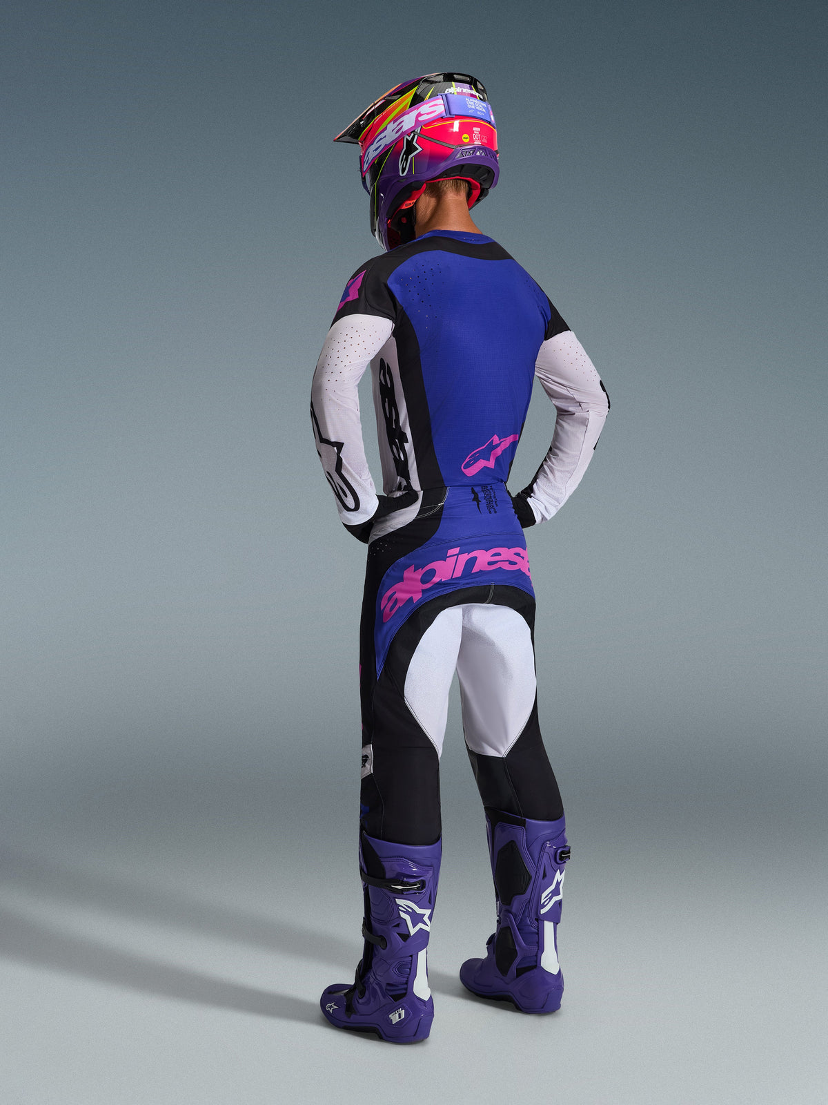 Motocrossrijder van achteren gezien in de Alpinestars 2026 Techstar Knif Gear Set, blauwe jersey met witte geperforeerde mouwen en roze logo's, zwart-witte broek met roze branding op de achterzijde, gecombineerd met paarse laarzen en een opvallende paarse en roze helm, professionele motorcross race-outfit.