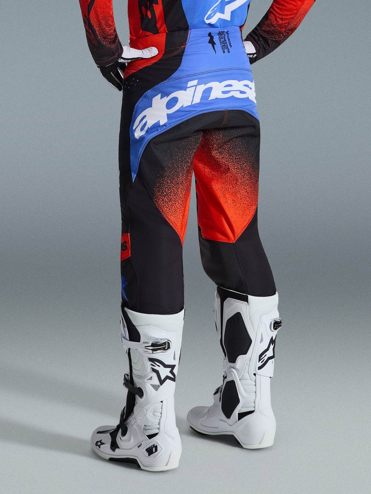 Alpinestars 2026 Techstar Knif Pants, Motocross Pants, Orange Black and UCLA Blue, rear view showing blue back yoke with white Alpinestars logo, orange gradient pattern on legs, worn with matching jersey and white boots for off-road racing.