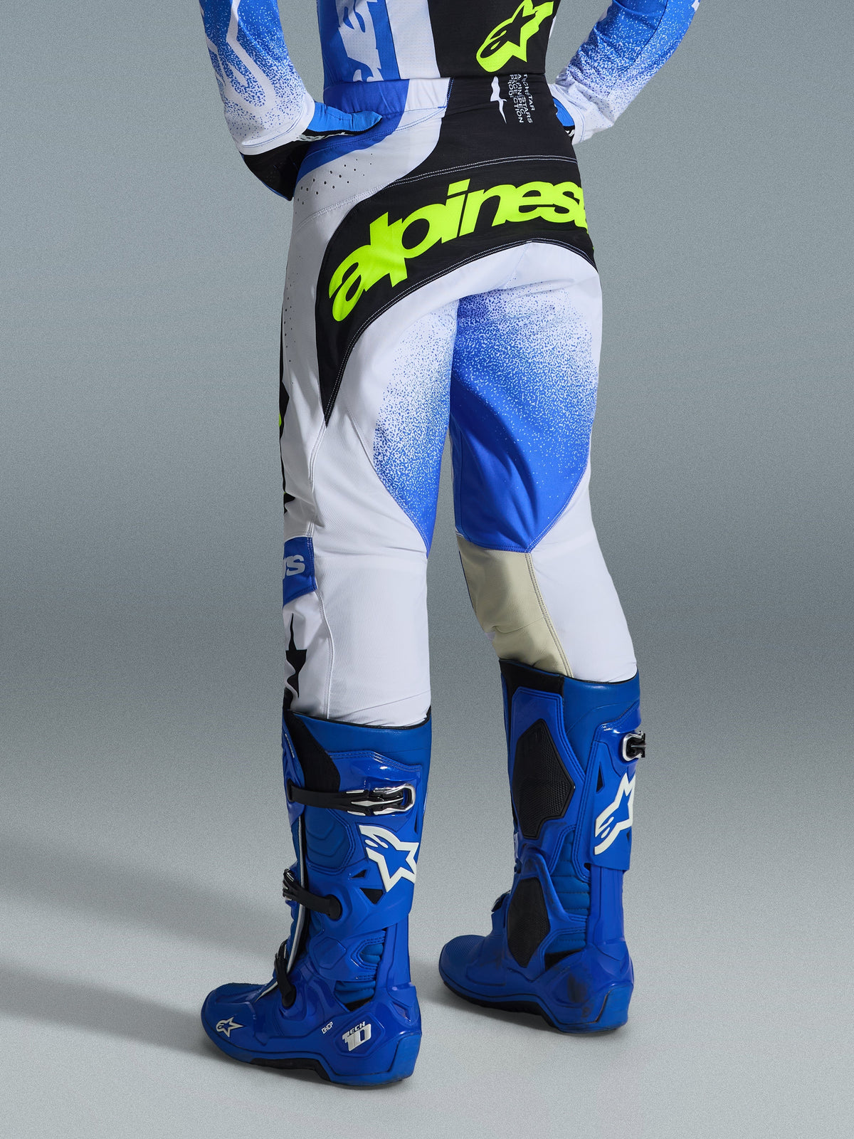 Alpinestars 2026 Techstar Knif broek, motorcrossbroek, Ucla Blue Black en Yellow Fluo, achteraanzicht met witte panelen en een blauw kleurverloop-spuitpatroon, opvallende geel-fluo branding op het zwarte zitvlak, gedragen met bijpassende blauwe Tech 10 laarzen en jersey voor off-road racen.
