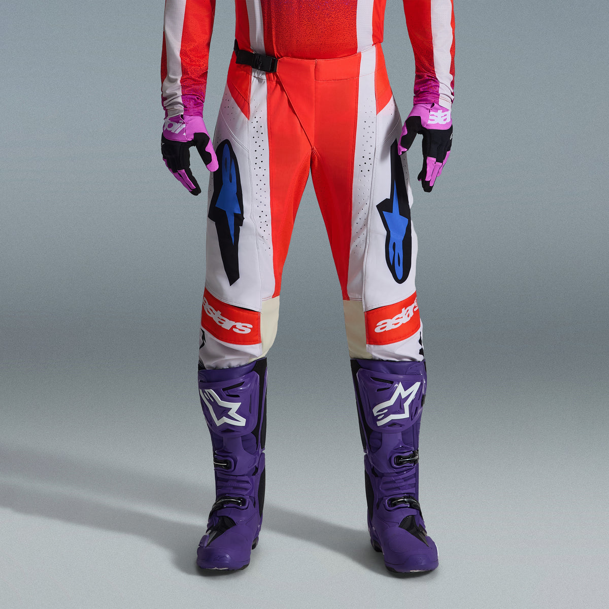 Alpinestars 2026 Techstar Knif Gear Set, onderlichaam van een motorcrosser die een rood-witte broek met blauwe accenten draagt, felpaarse racelaarzen met witte logo's en bijpassende zwart-paarse handschoenen, professionele off-road race-uitrusting