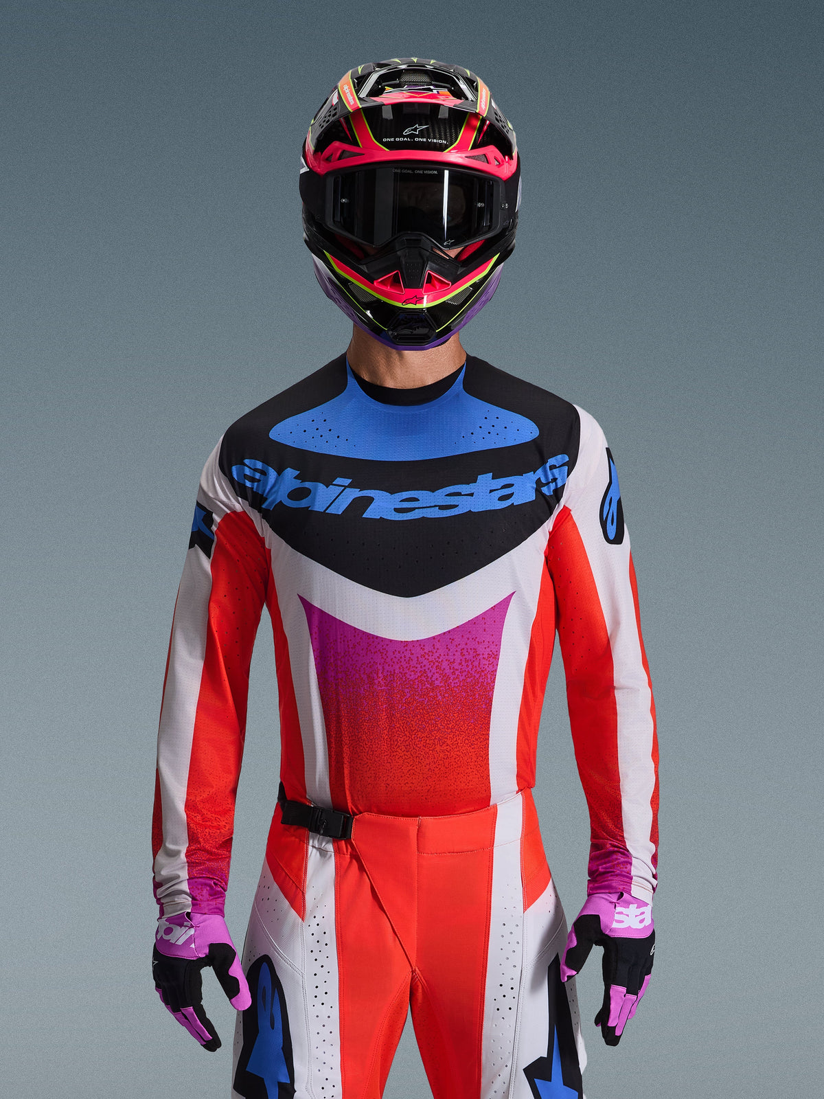 Motorcrossrijder die de Alpinestars 2026 Techstar Knif Jersey draagt in Light Gray Orange Fluo Black, voorzien van een opvallend blauw logo en feloranje mouwen, gedragen met een roze en zwarte helm en paarse handschoenen, atletische racepasvorm voor hoogwaardige off-road competities