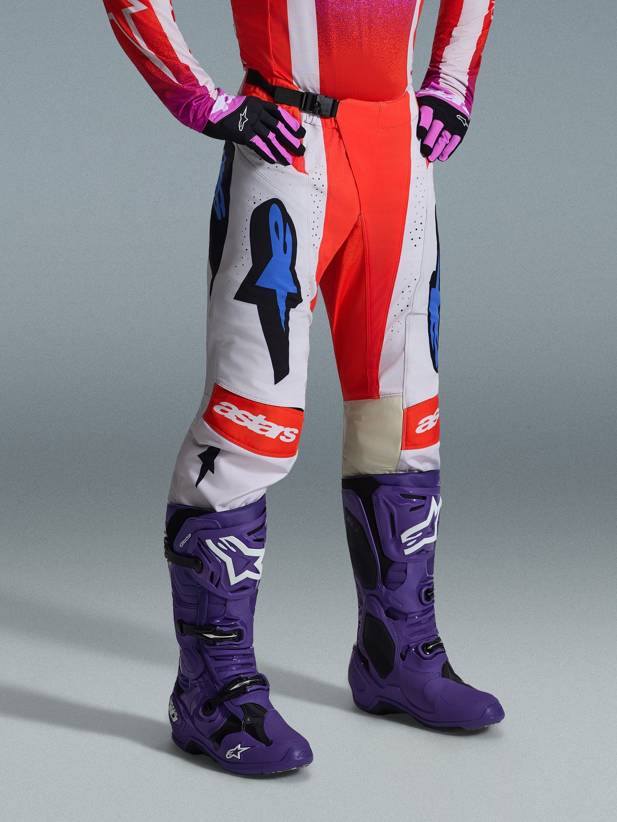 Alpinestars 2026 Techstar Knif broek, motorcrossbroek, wit en oranje met blauwe accenten, gedragen als onderdeel van een complete uitrusting met een rood en roze shirt, paarse laarzen en roze handschoenen, voorzien van opvallende logo's en een atletische racepasvorm