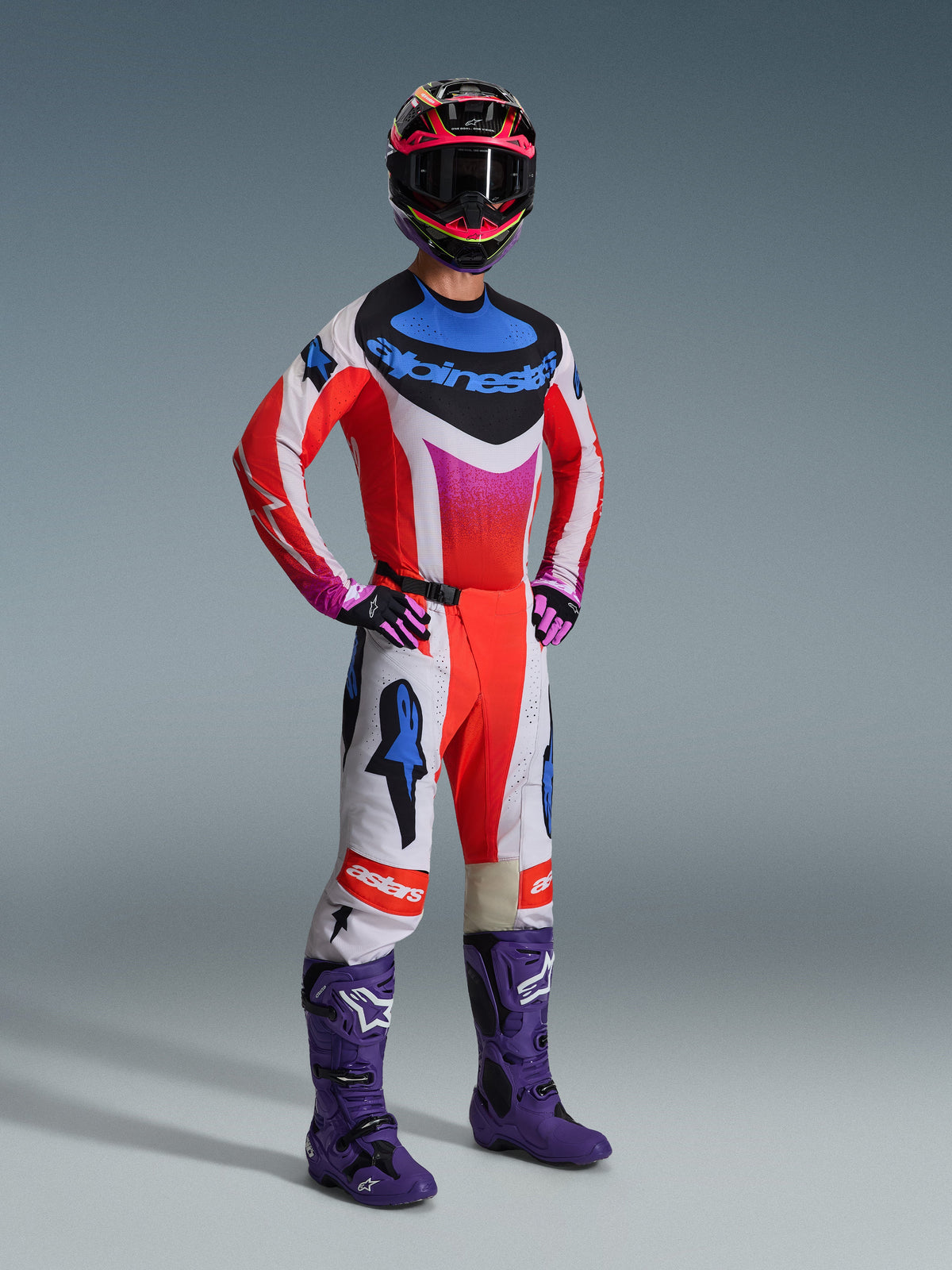 Alpinestars motorcrossrijder in volledige race-uitrusting, 2026 Techstar Knif Jersey in wit en rood met blauwe en roze accenten, bijpassende motorcrossbroek, paarse laarzen en een veelkleurige helm, professionele outfit voor off-road prestaties