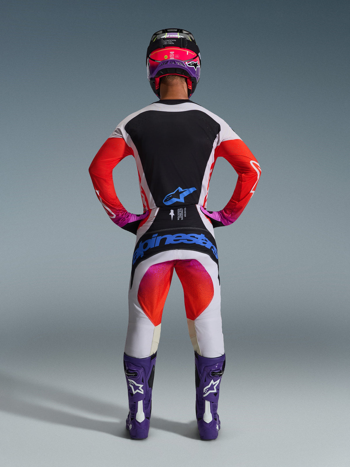 Alpinestars 2026 Techstar Knif race-uitrusting, motorcrossrijder van achteren gezien, lichtgrijs shirt met zwart rugpaneel en oranje mouwen, bijpassende witte en oranje broek met blauwe Alpinestars-branding, paarse laarzen en een zwart-roze helm, gecoördineerde professionele outfit voor off-road racing.