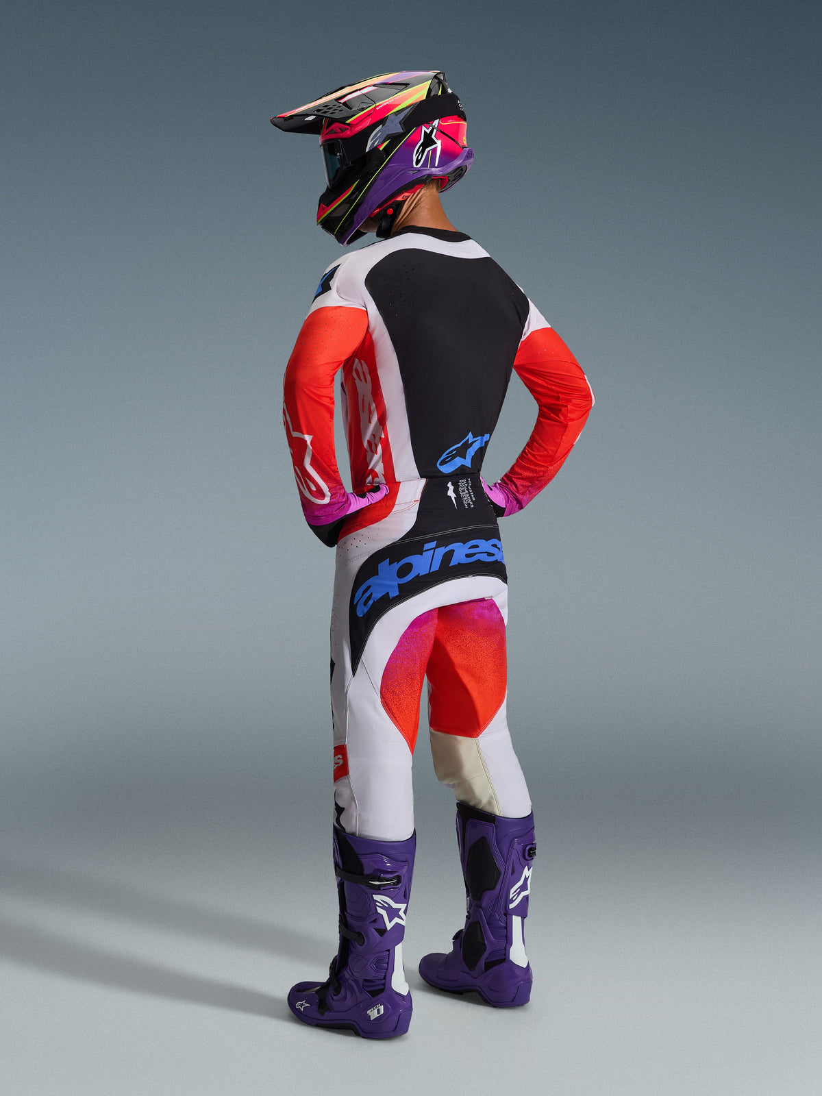 Alpinestars 2026 Techstar Knif Gear Set, motorcrosser van achteren gezien, wit shirt met rode mouwen en zwart paneel, witte broek met rode en paarse accenten en blauwe Alpinestars logo's, paarse racelaarzen en bijpassende helm, professionele off-road race-uitrusting